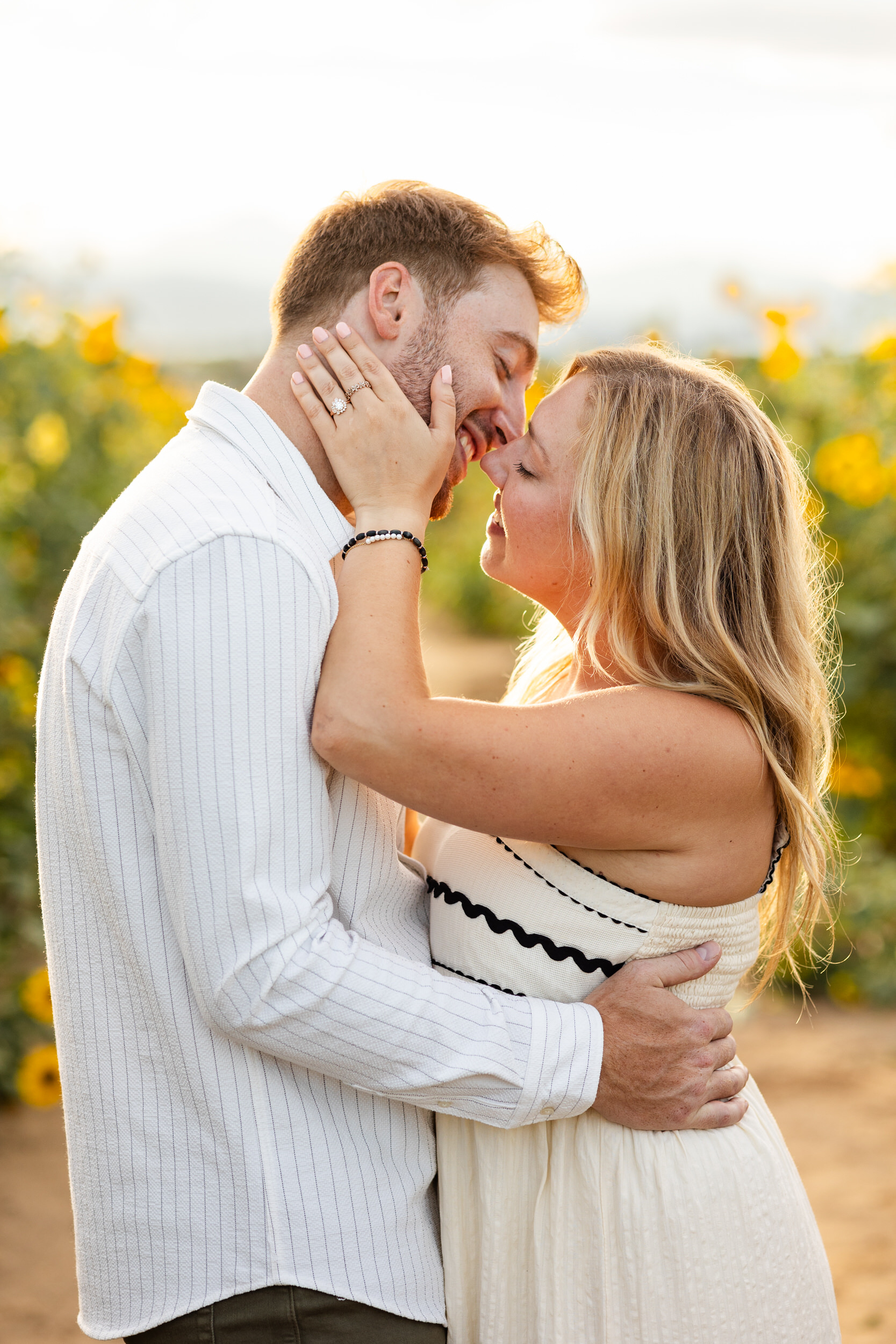 A newly engaged couple lean in for a kiss in a sunflower field.