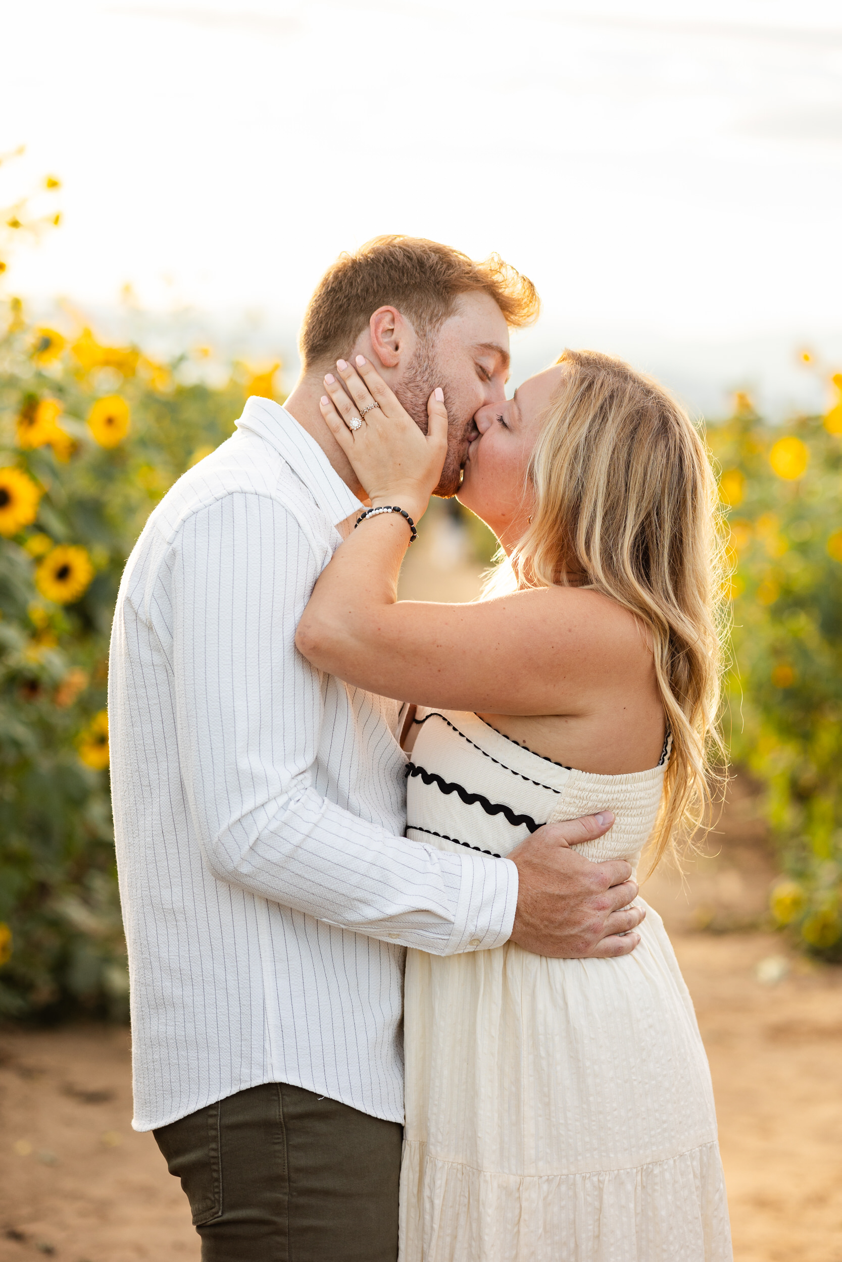 An engaged couple shares a kiss in a sunflower field.