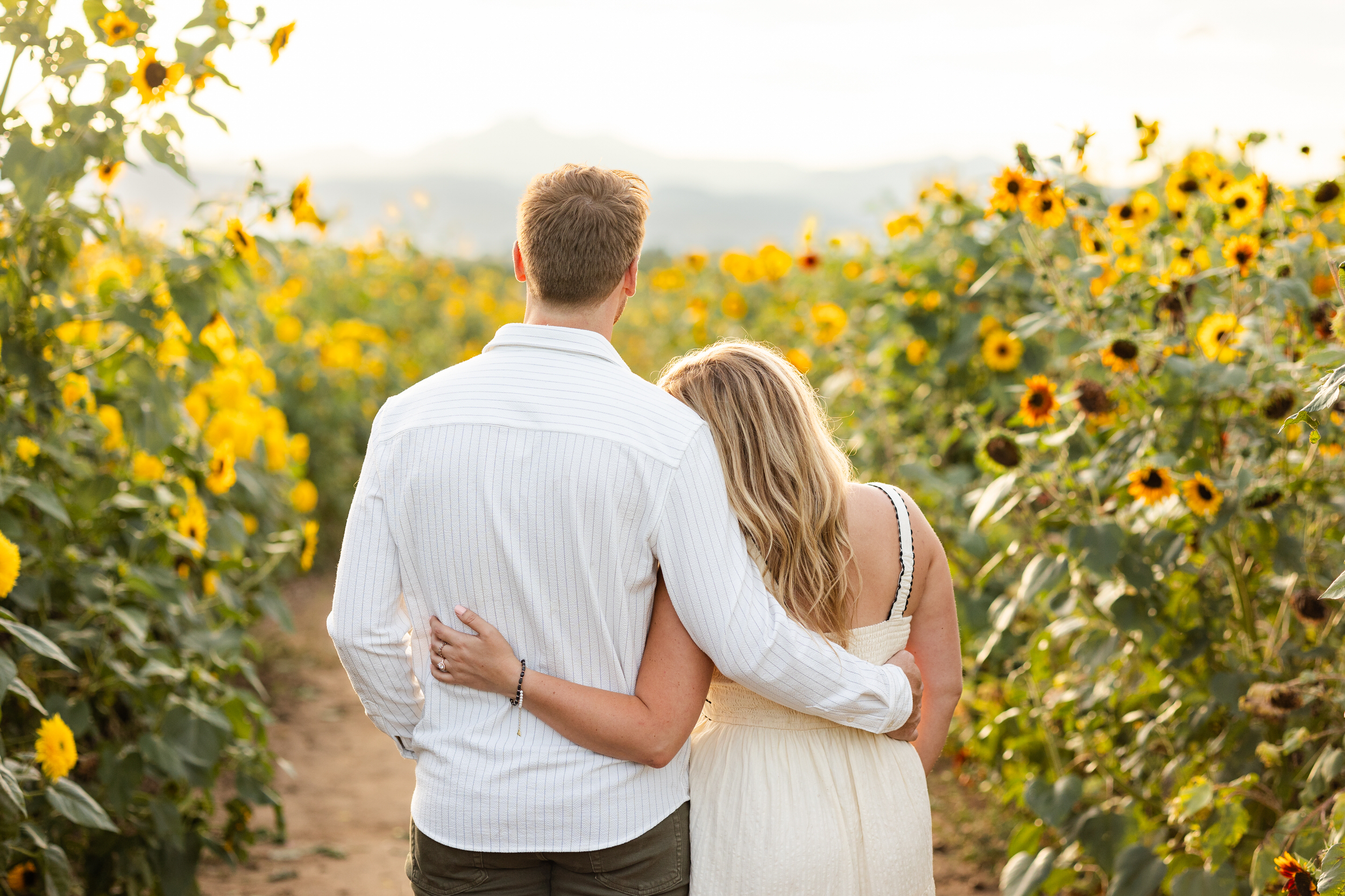 A newly engaged couple stands in a sunflower field with their arms around each other's shoulders.