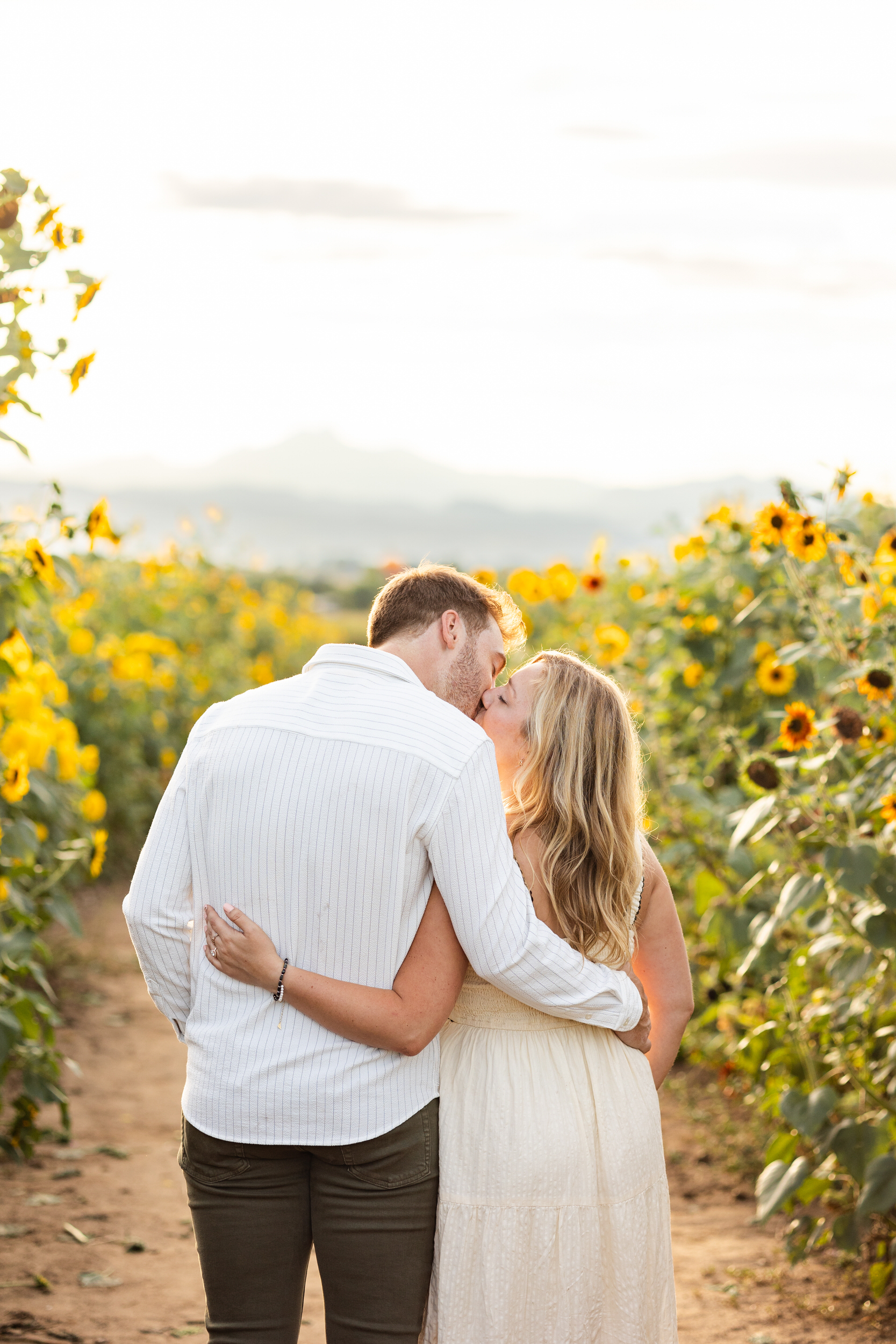 An engaged couple shares a kiss in a sunflower field.