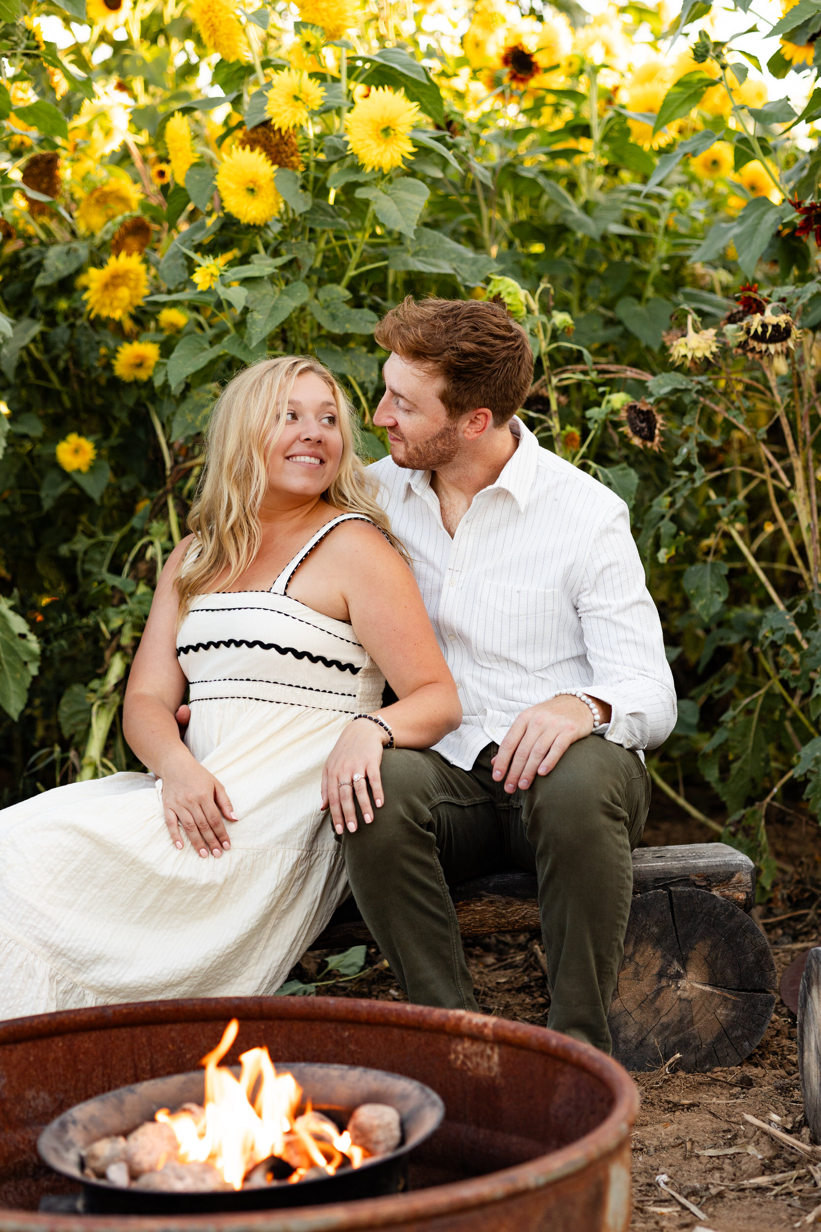A woman leans her back on her fiancé as they snuggle close around a campfire.