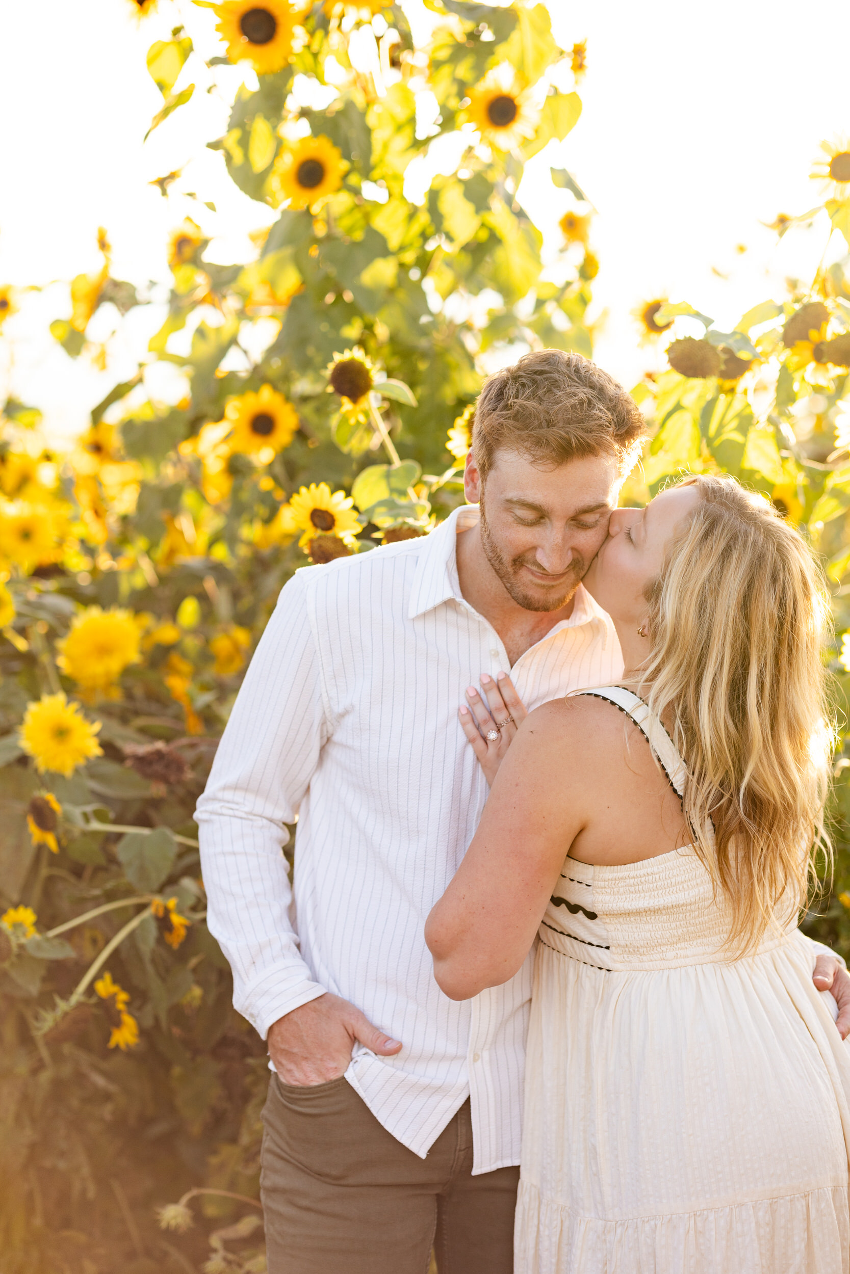 A woman kisses her fiancé's cheek in a sunflower field.