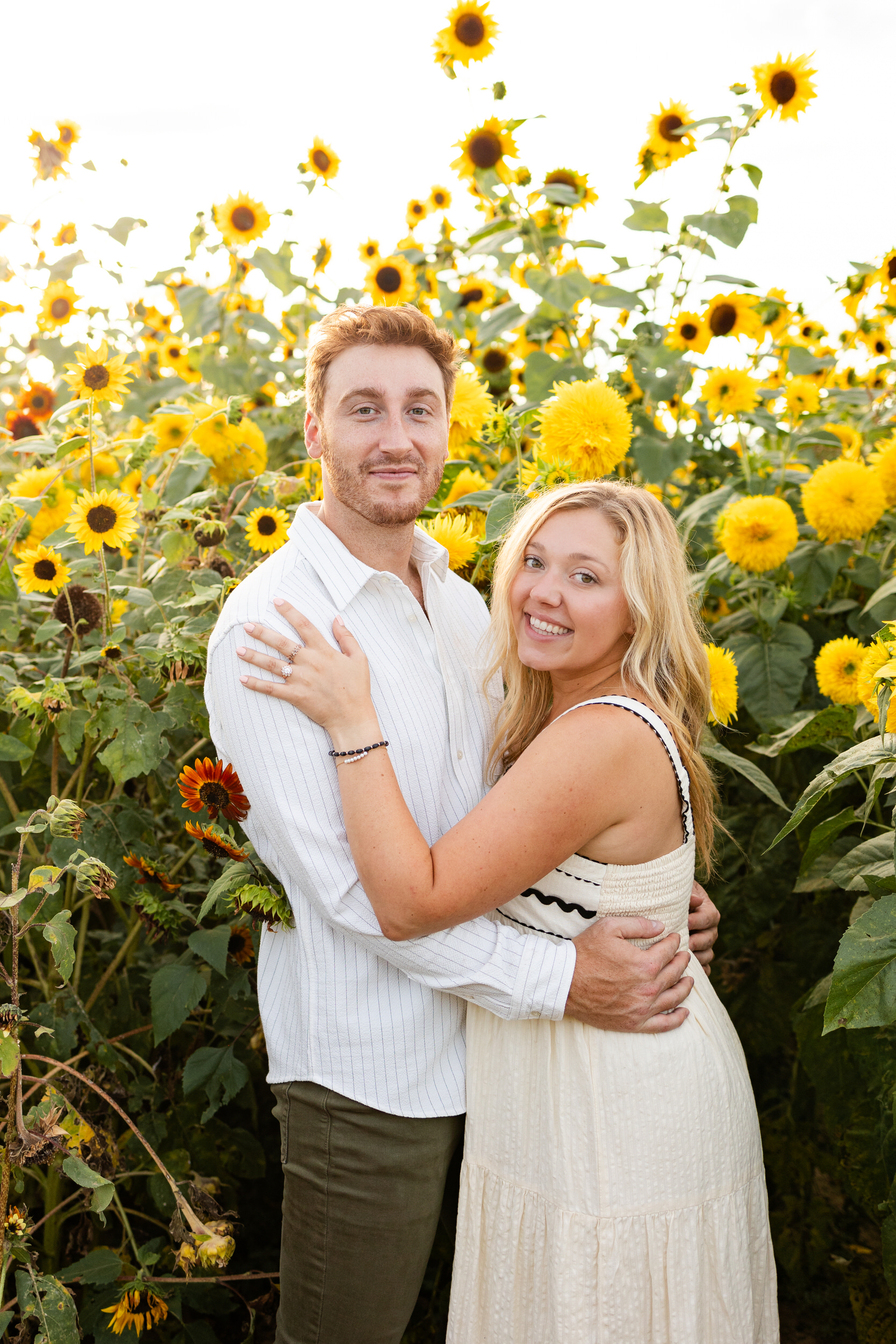 A newly engaged couple smiles at the camera in a sunflower field.