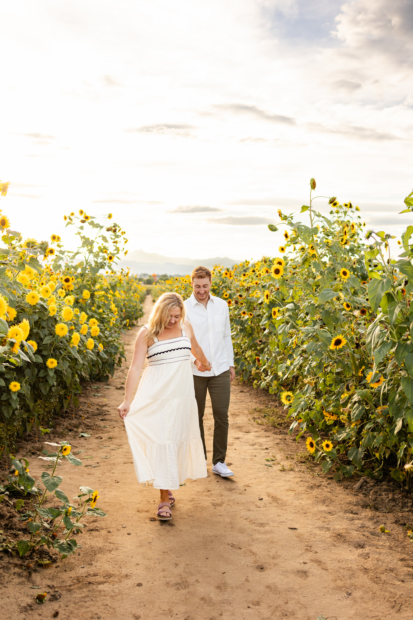 A woman holds her fiancée's hand and leads him through a sunflower field.