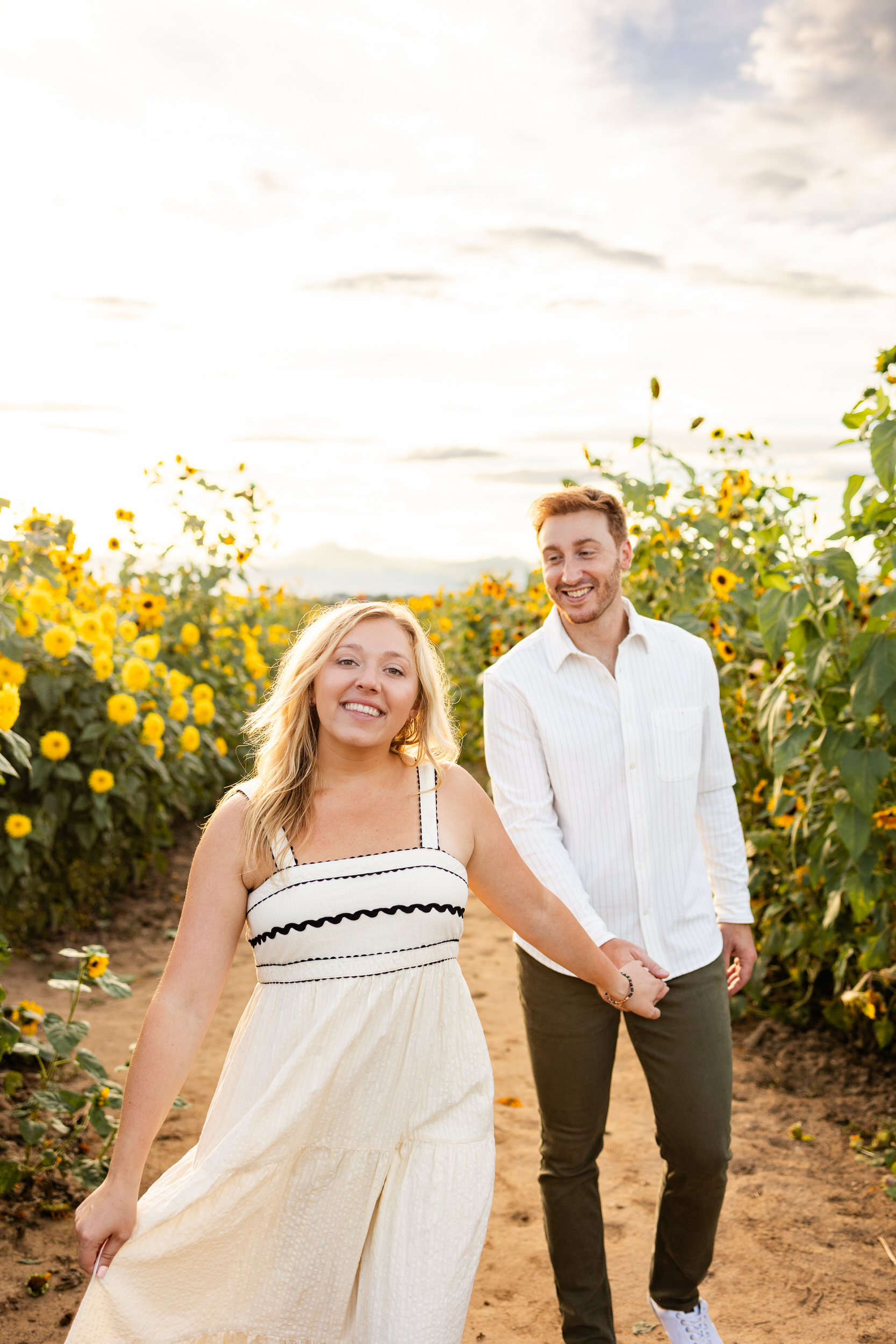 A woman holds her fiancée's hand and leads him through a sunflower field.