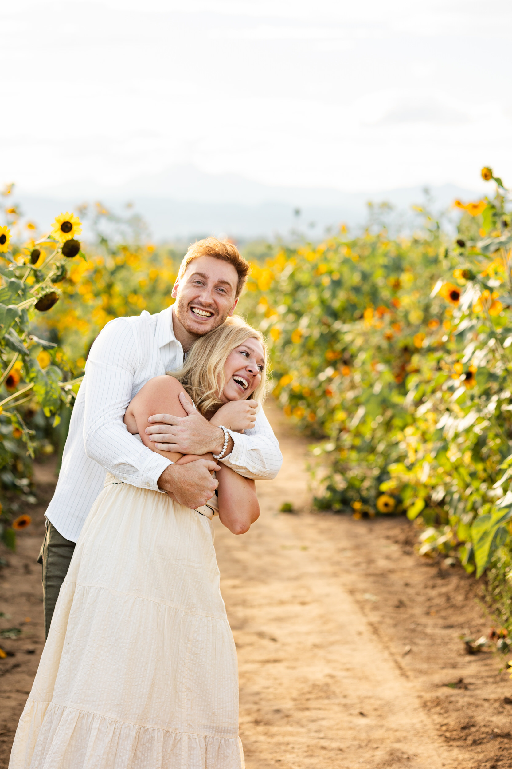 A man hugs his fiancée around her shoulders in a sunflower field as they laugh together.
