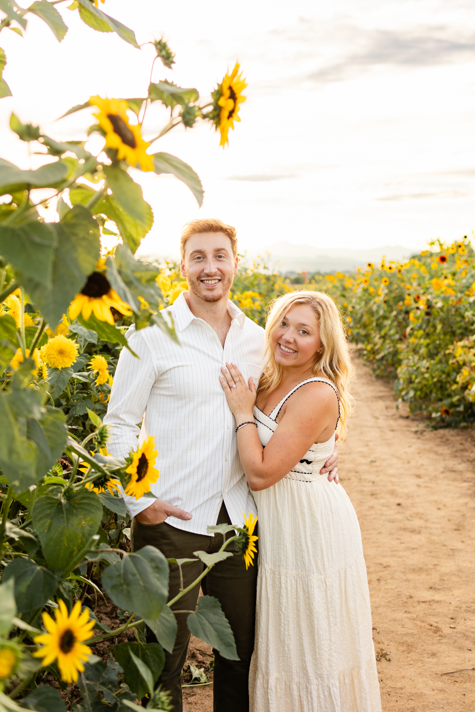 An engaged couple smiles at the camera in a sunflower field.