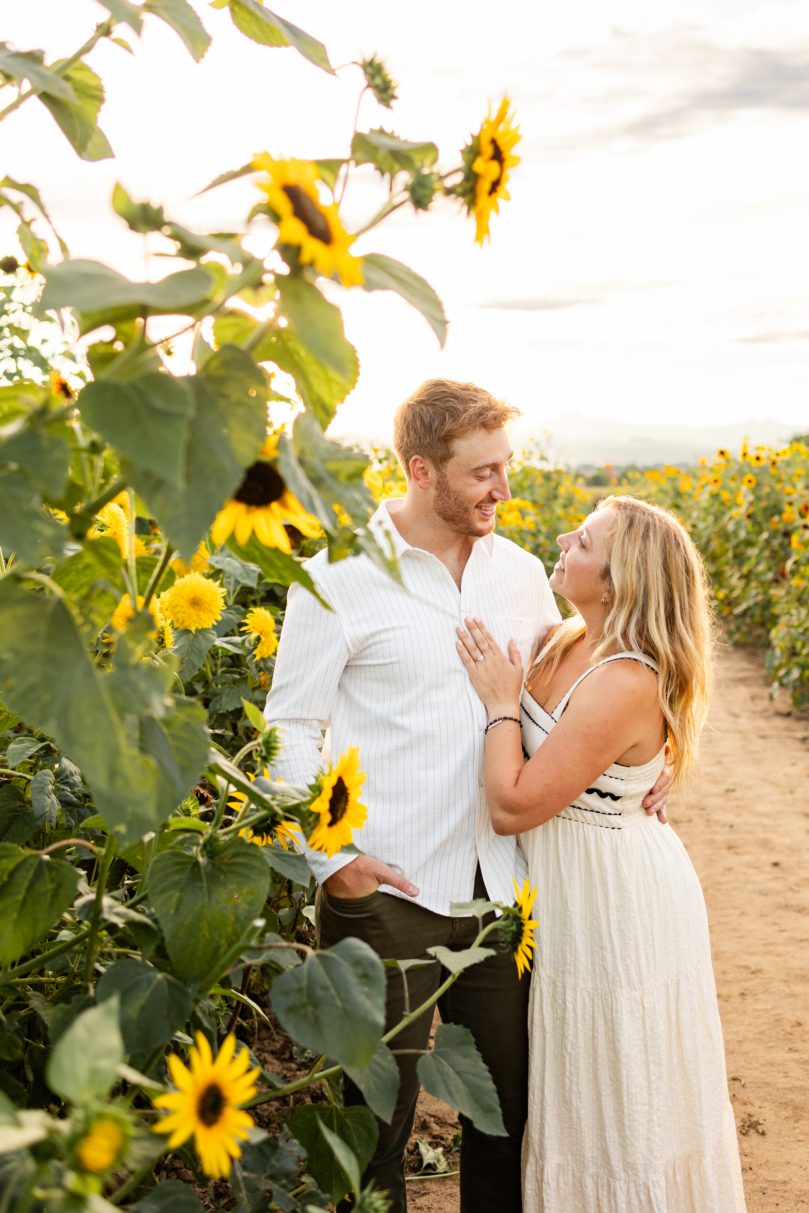 An engaged couple looks and smiles at each other in a sunflower field.