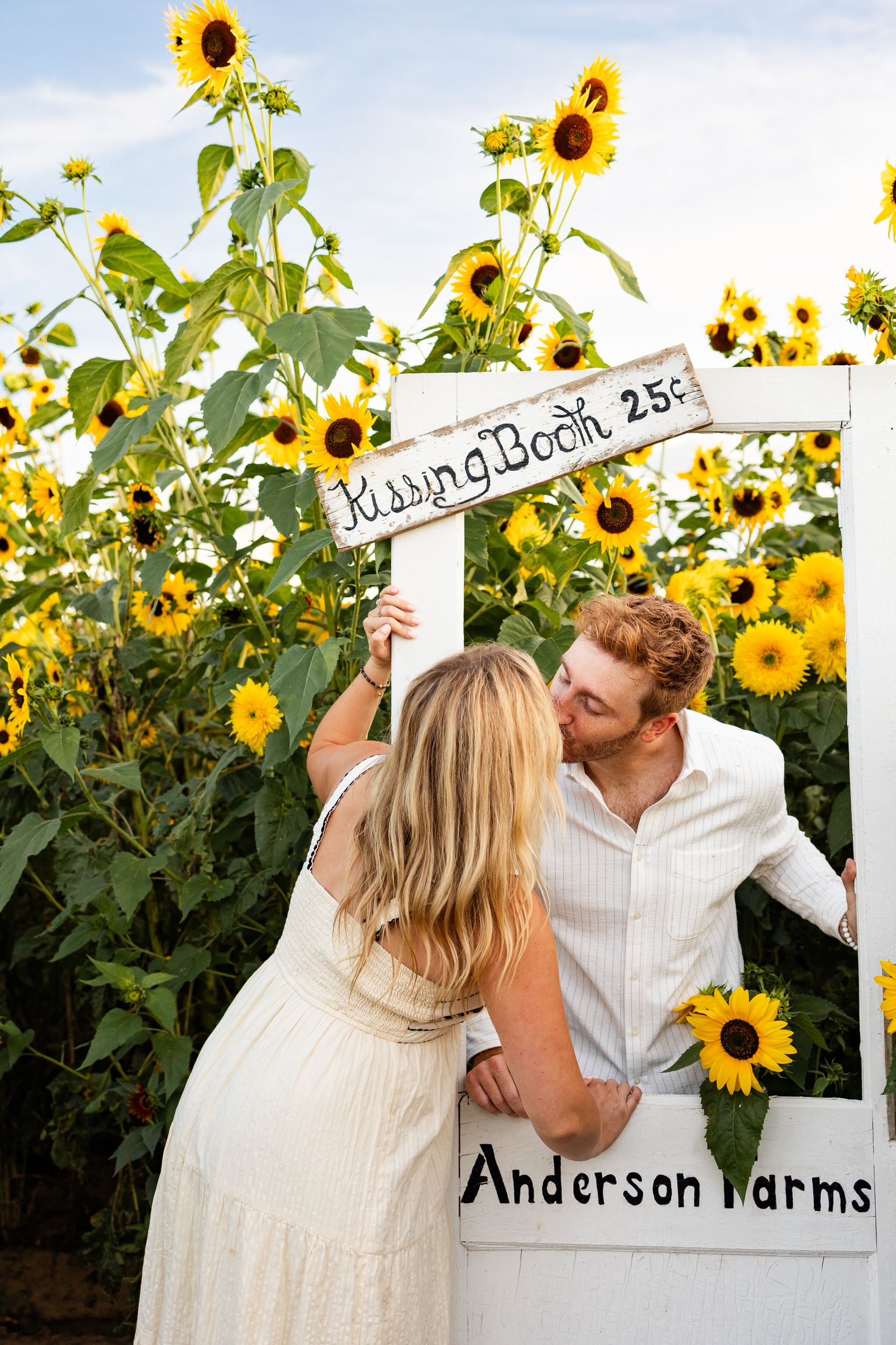 A newly engaged couple shares a kiss at a kissing booth in a sunflower field.