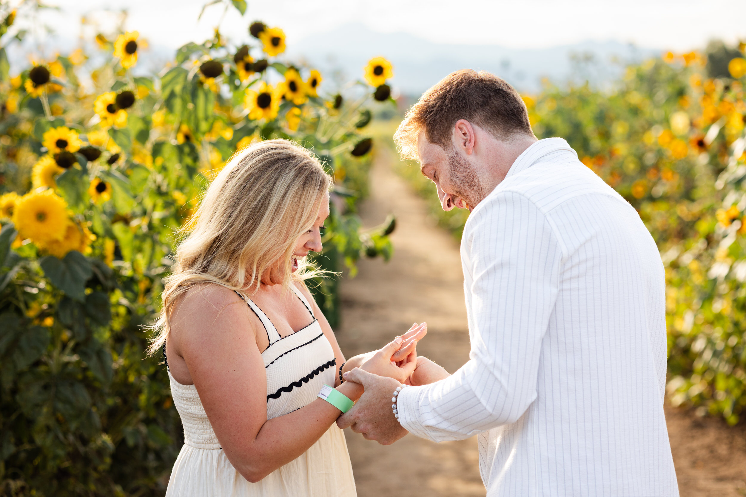 A woman looks at her ring with a big smile as her fiancé puts the ring on her finger.