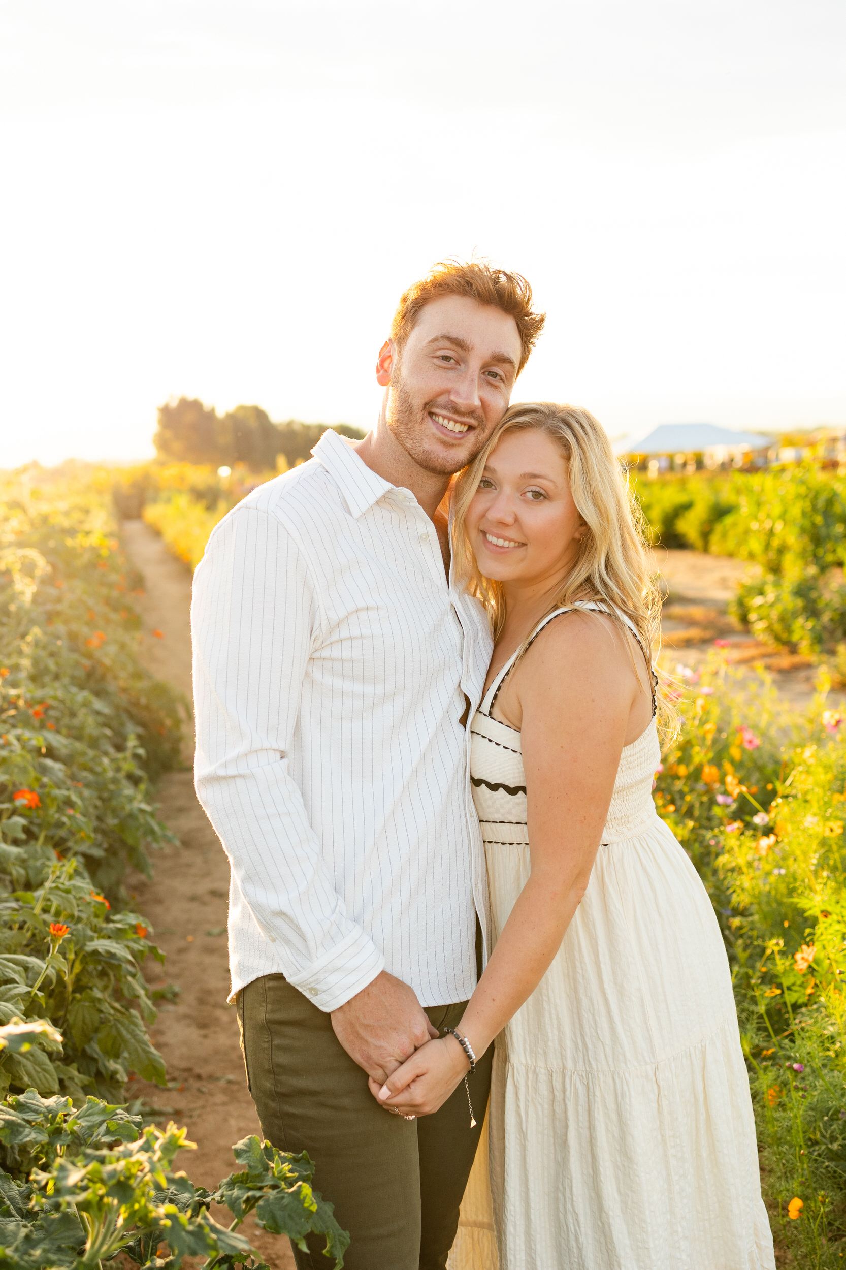 A newly engaged couple smiles at the camera in a flower field at sunset.