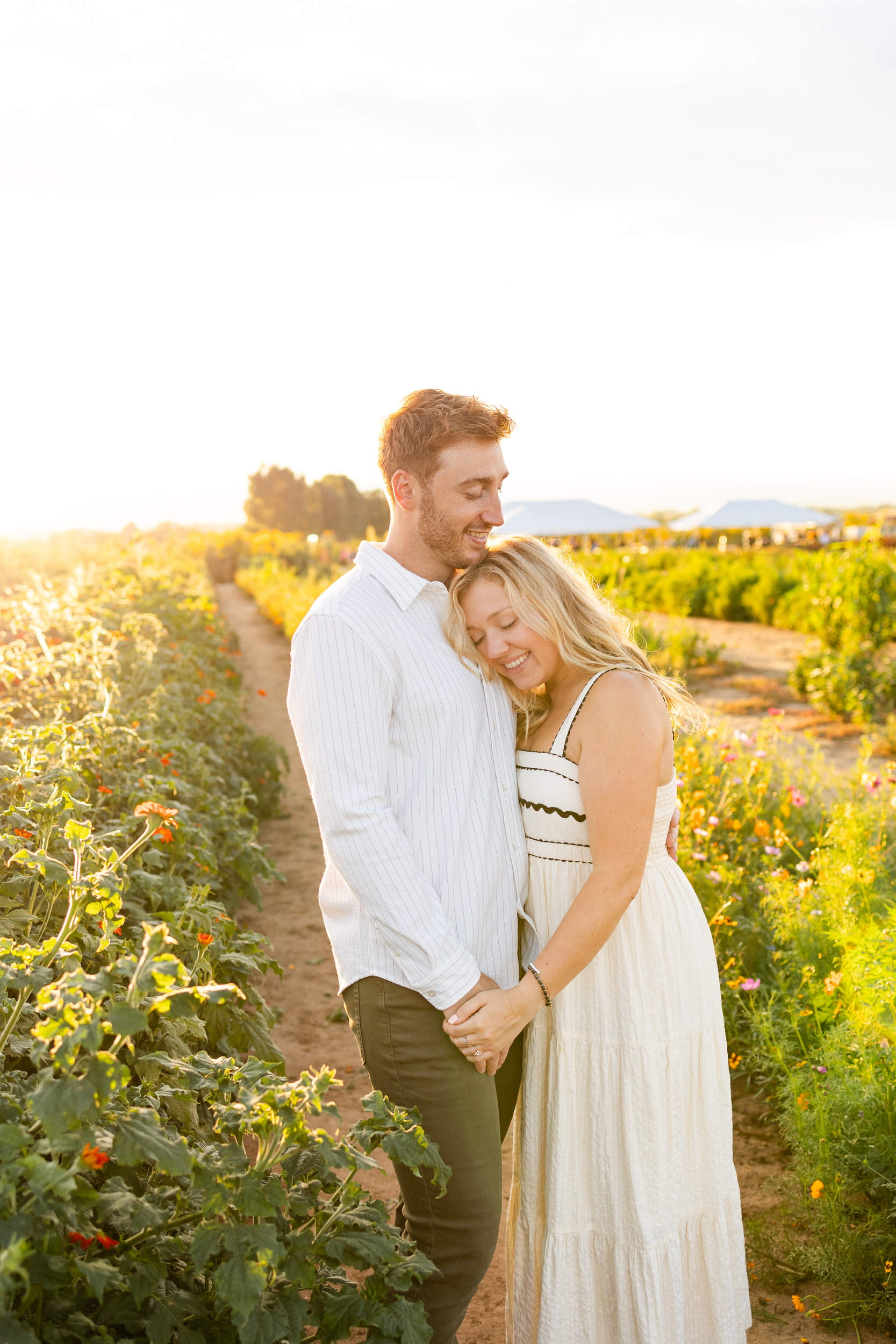 A woman leans her head on her fiancé's chest in a field of flowers at sunset.