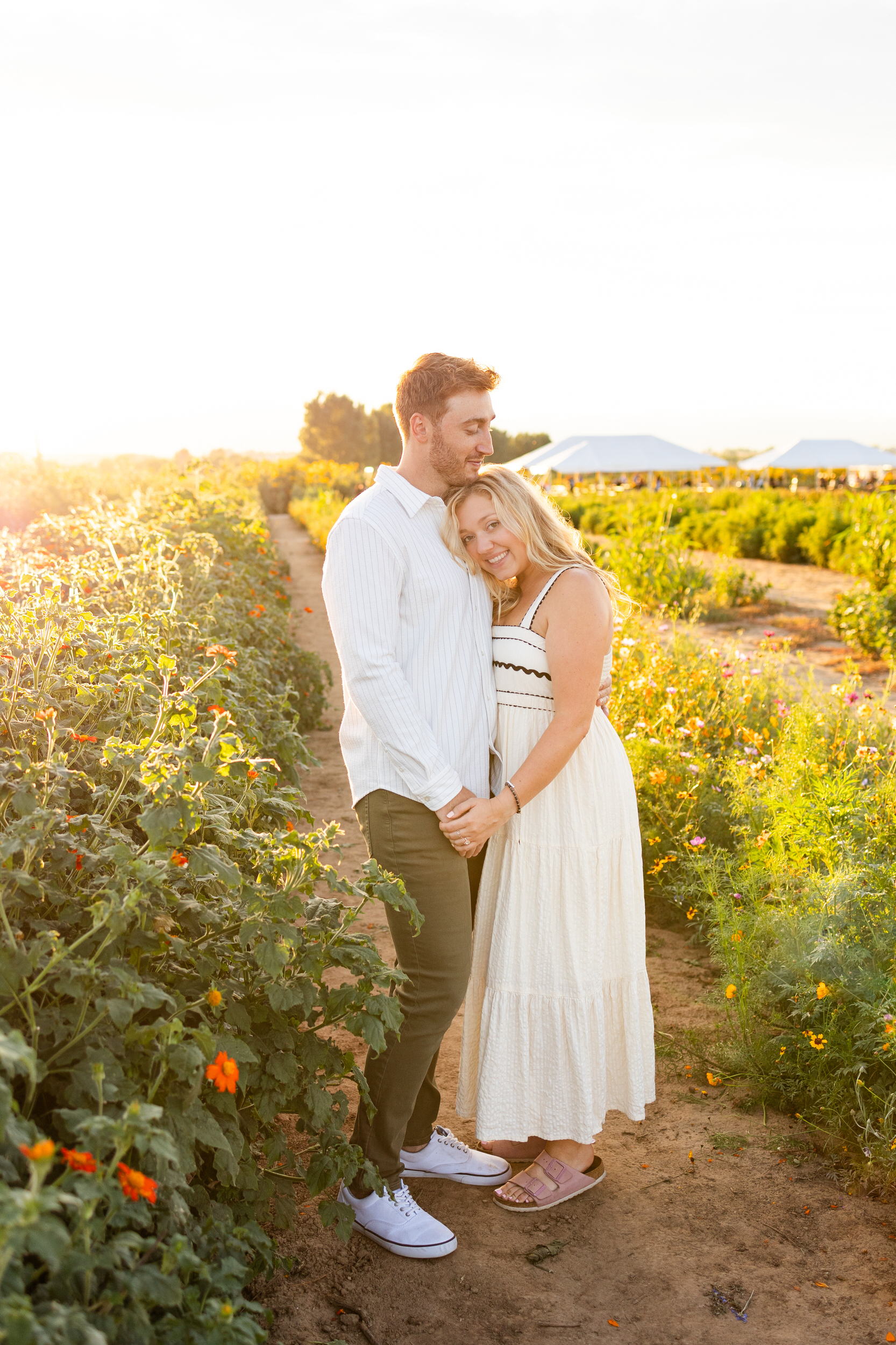 A woman leans her head on her fiancé's chest in a field of flowers at sunset.