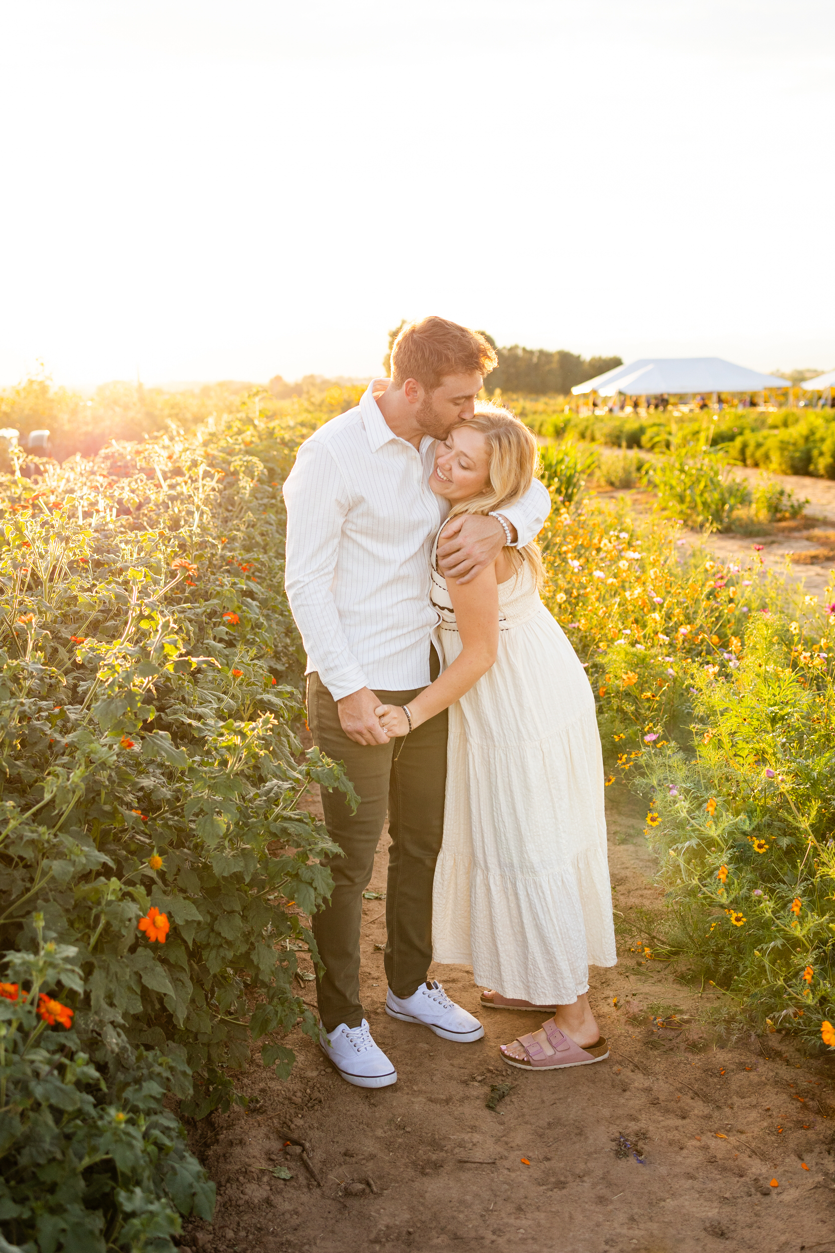 A man kisses the top of his fiancé's head as she leans her head on his chest in a field of flowers at sunset.