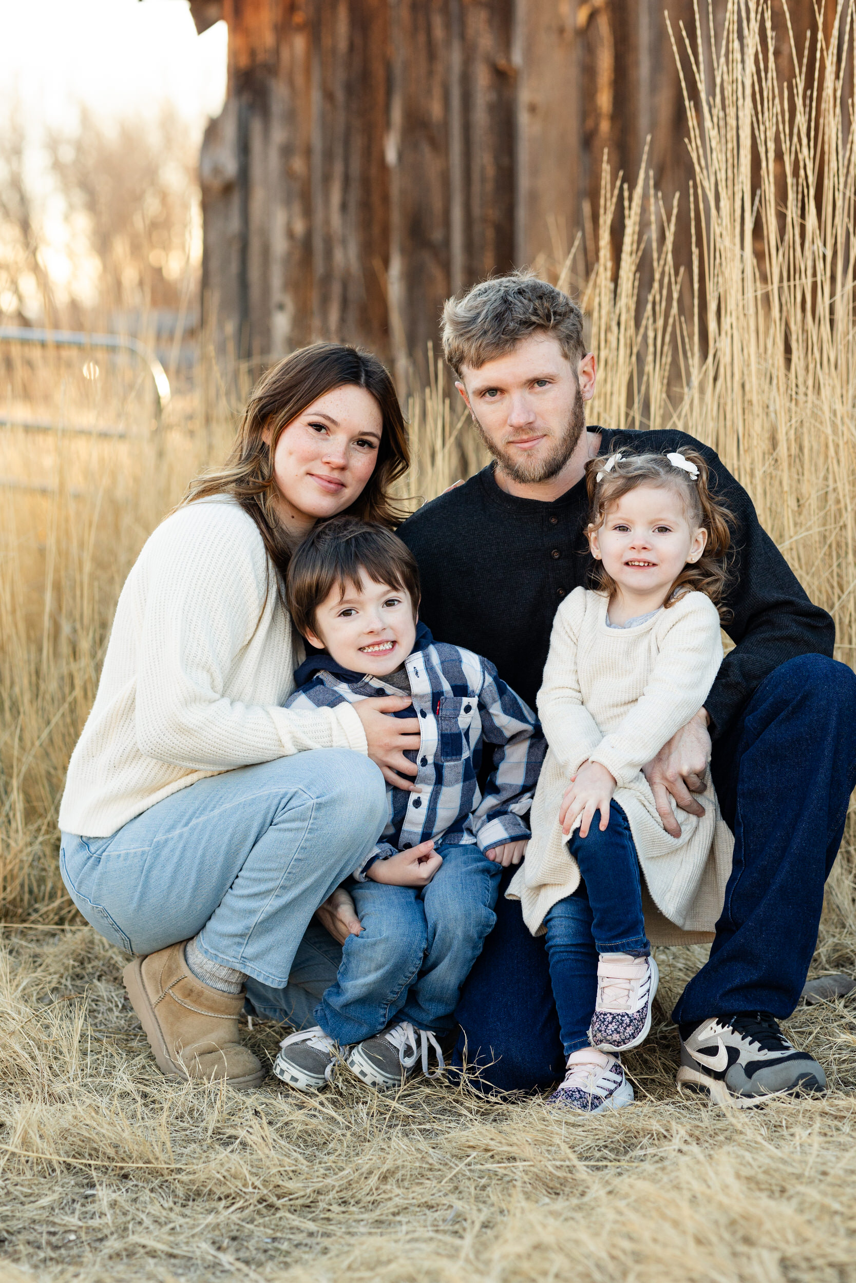 A mom and dad kneel next to their young son and daughter and they all smile at the camera.