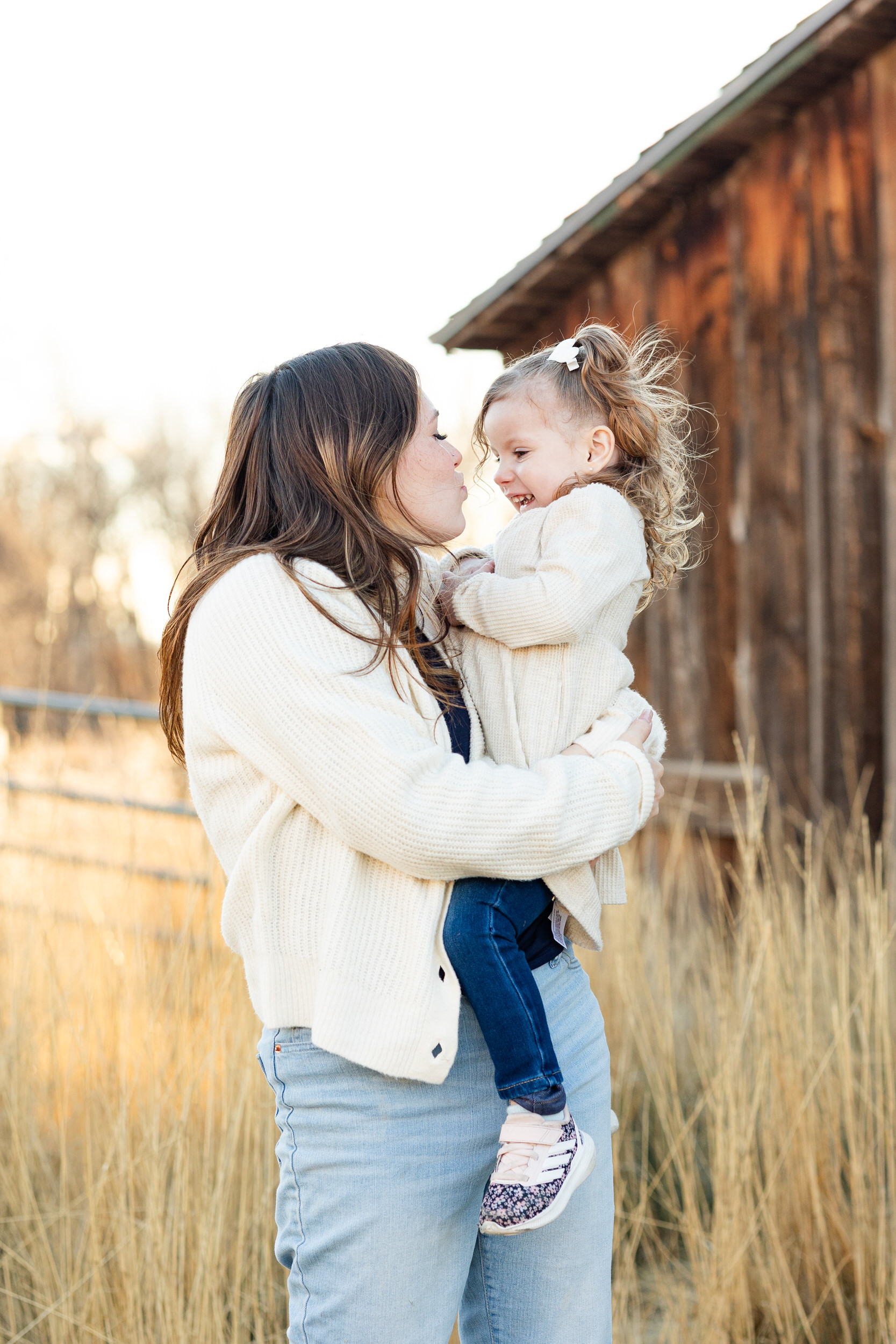 A mom holds her young daughter on her hip and tries to kiss her cheek as her daughter laughs.