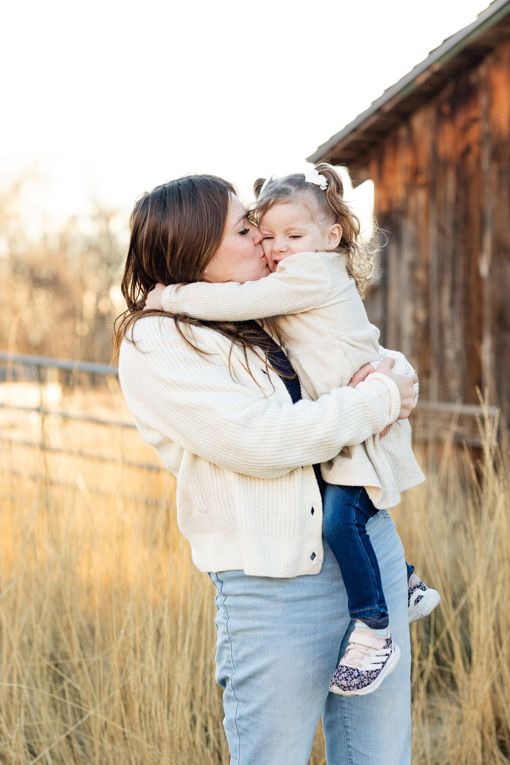 A mom holds her young daughter on her hip and kisses her cheek.