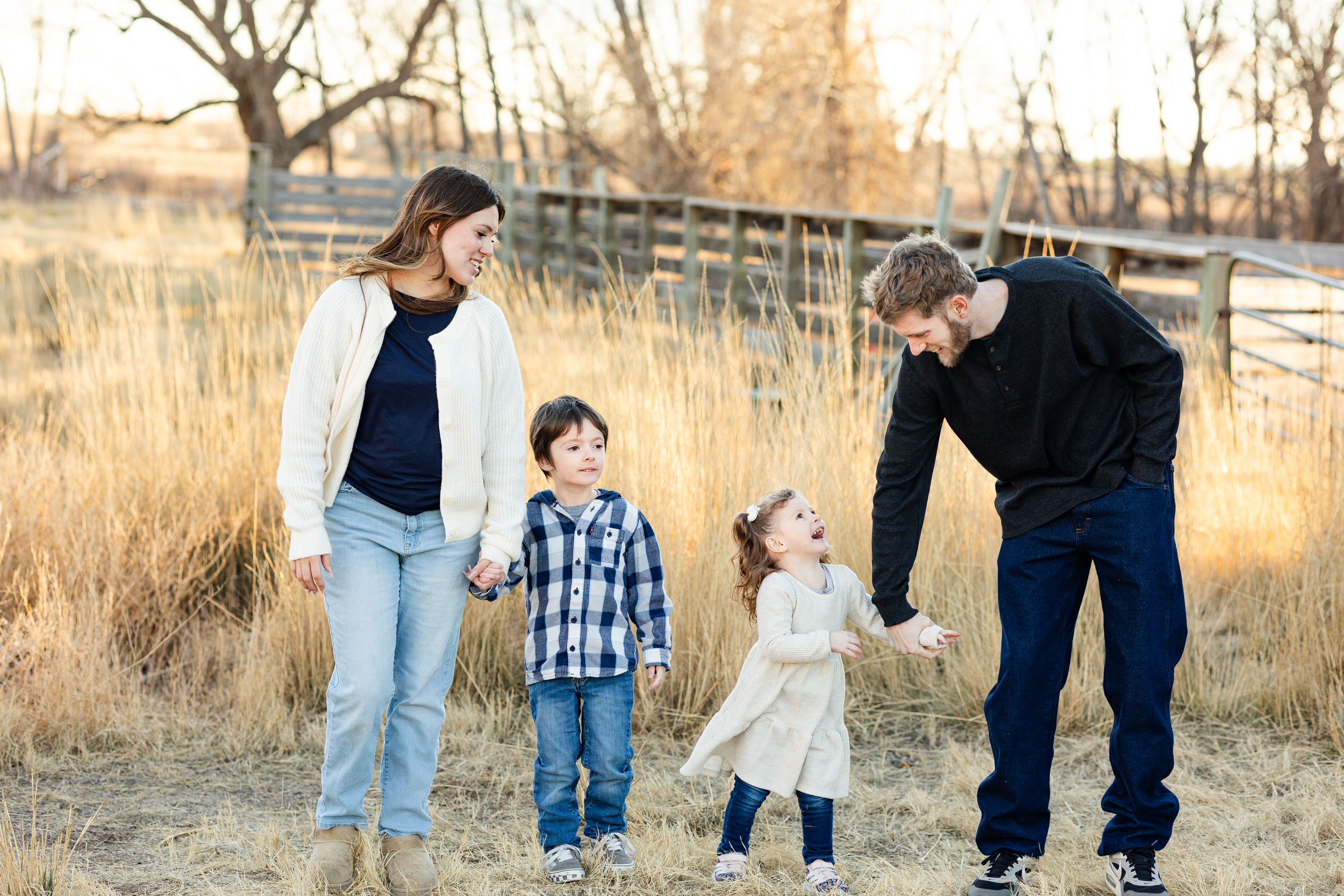 A mom and dad walk through a field with their young son and daughter while they all smile at each other.