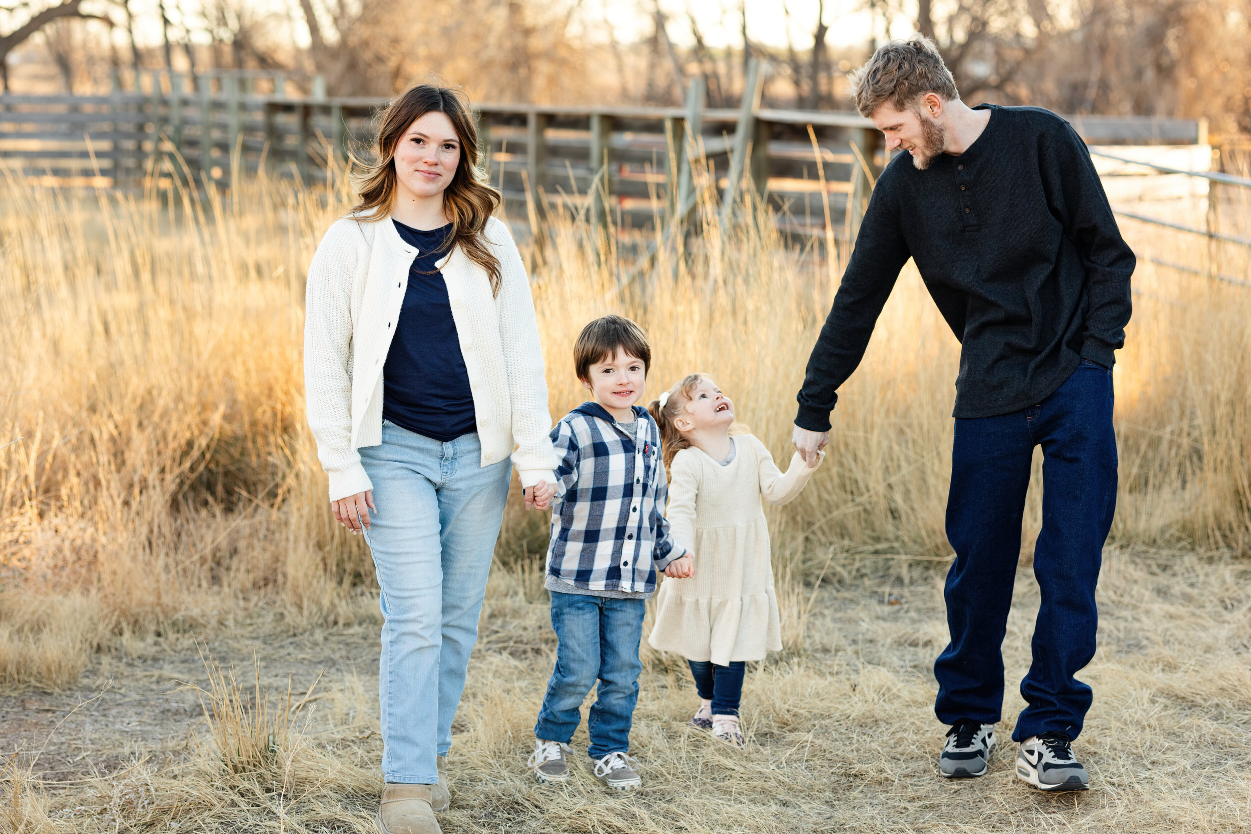A mom and dad walk through a field with their young son and daughter while they all smile at each other and the camera.