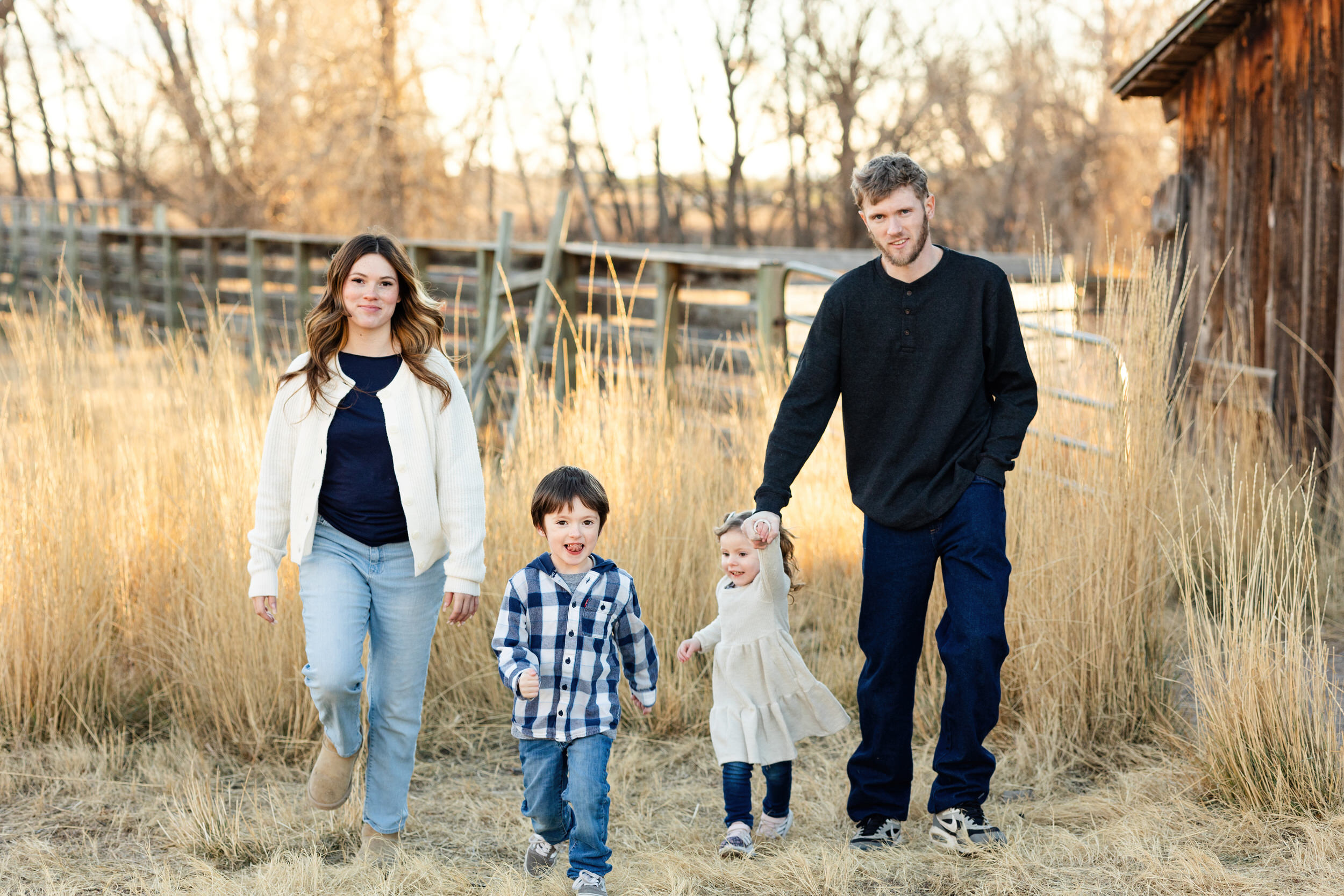 A mom and dad walk through a field with their young son and daughter while they all smile at each other and the camera.