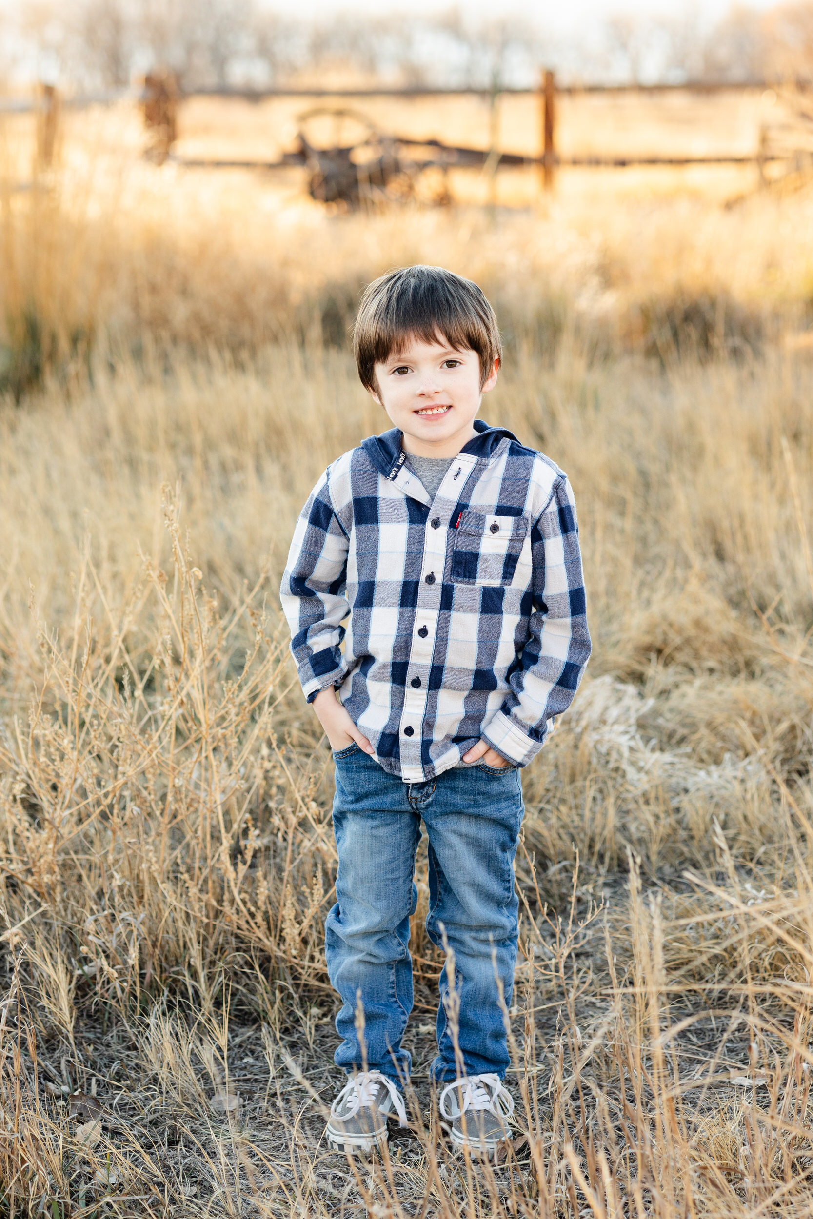A young boy stands with his hands in pockets and smiles at the camera.
