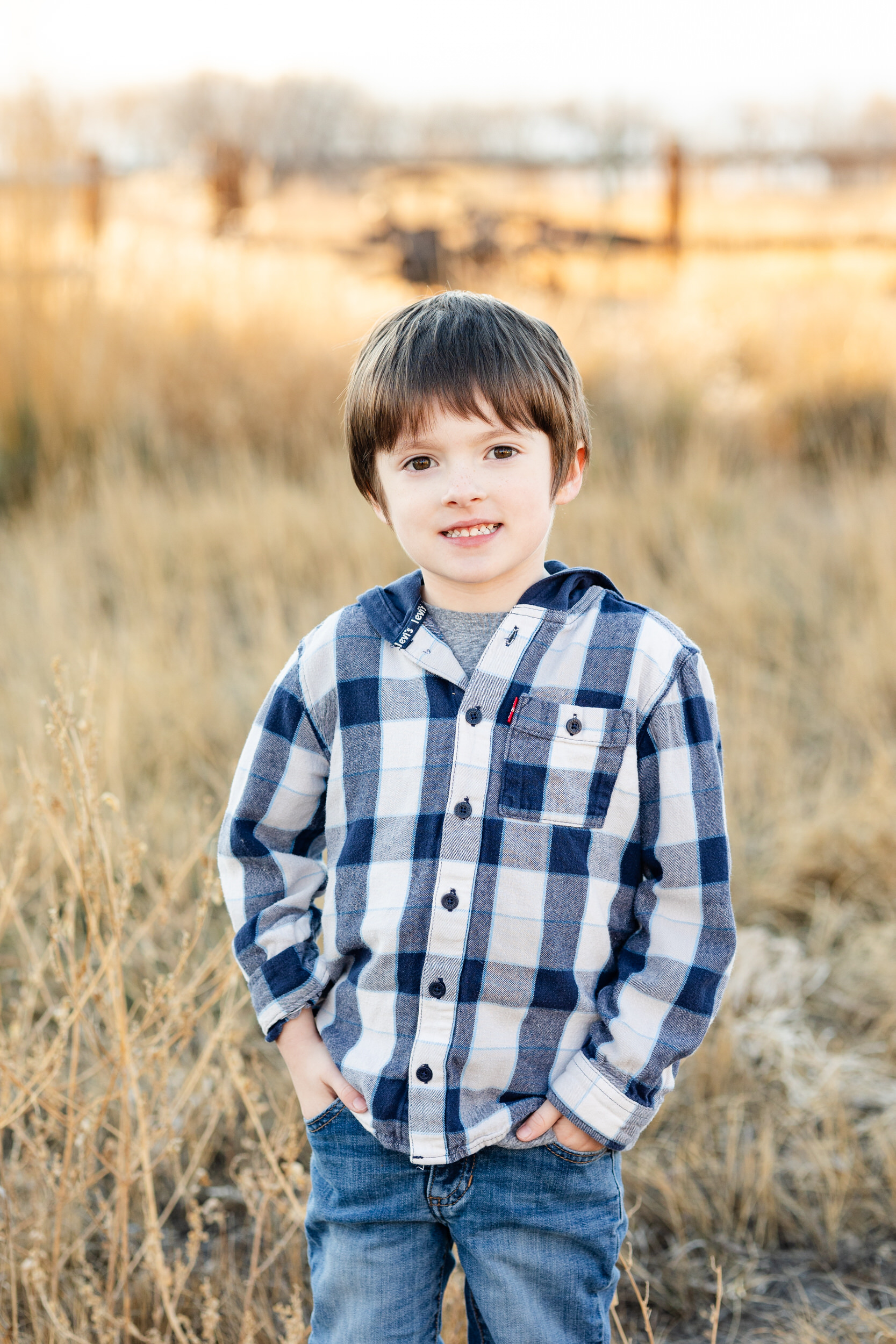A young boy stands with hands in pockets and smiles at the camera.