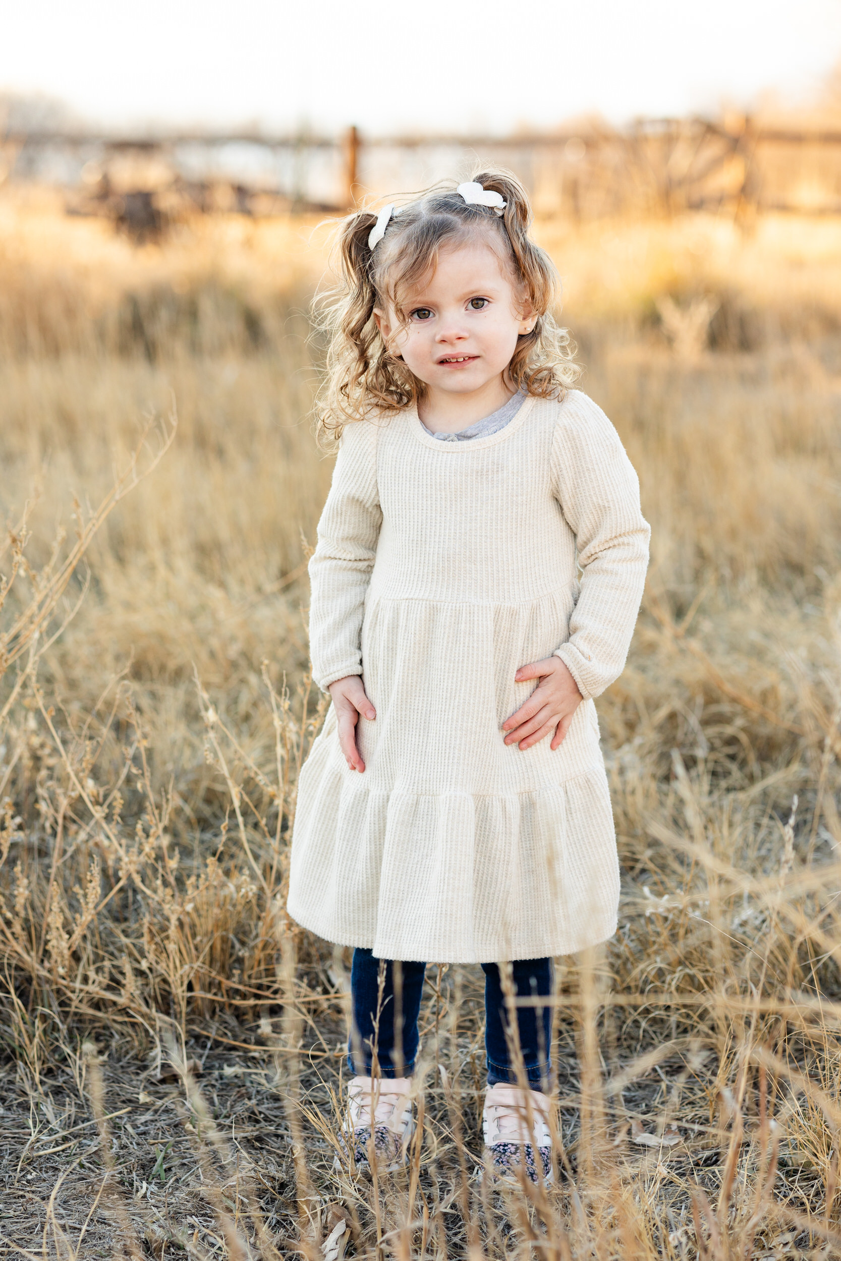 A little girl in a white dress stands in a field and looks at the camera.