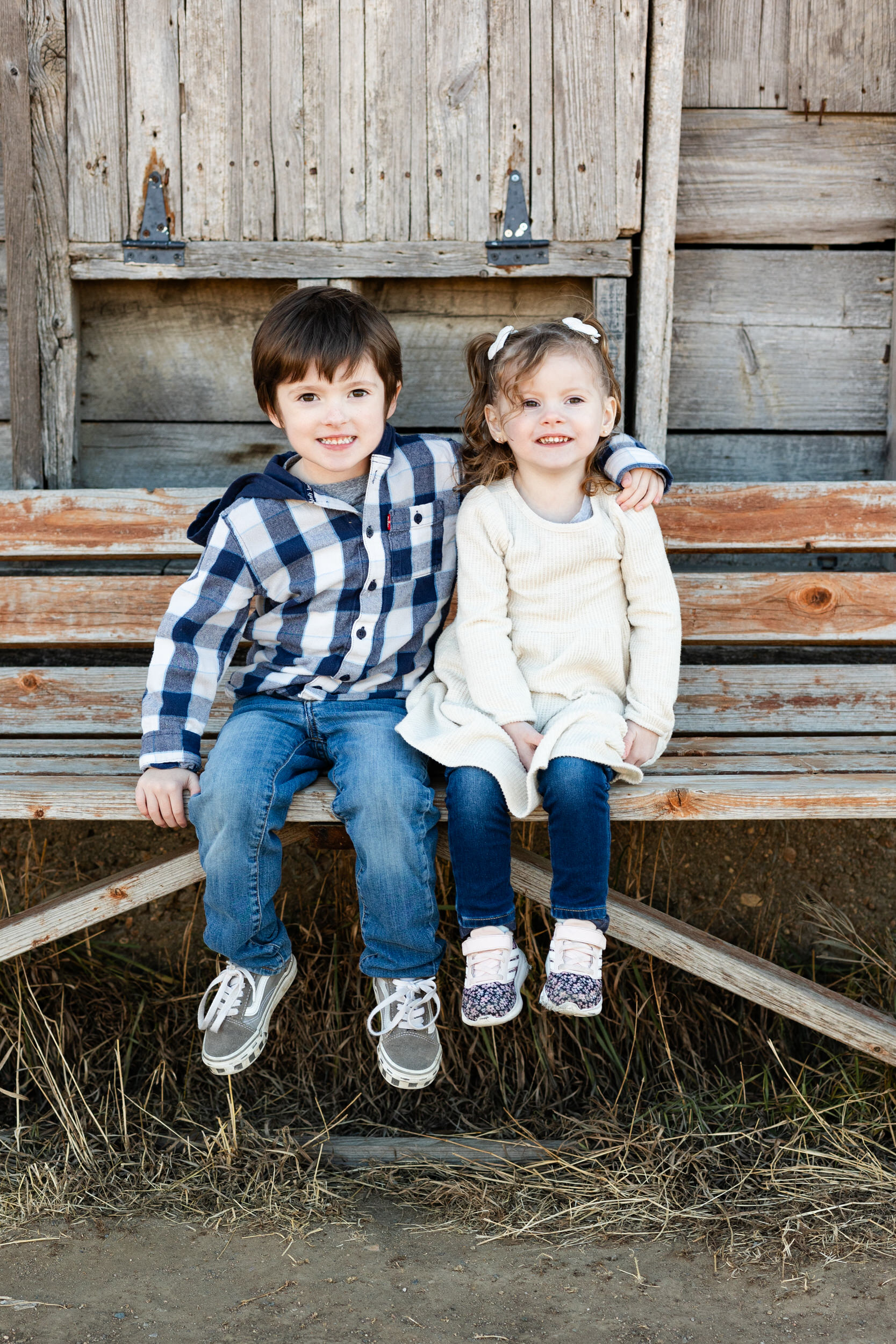 A little boy and girl sit on a bench with their arms around each other's shoulders and smile at the camera.