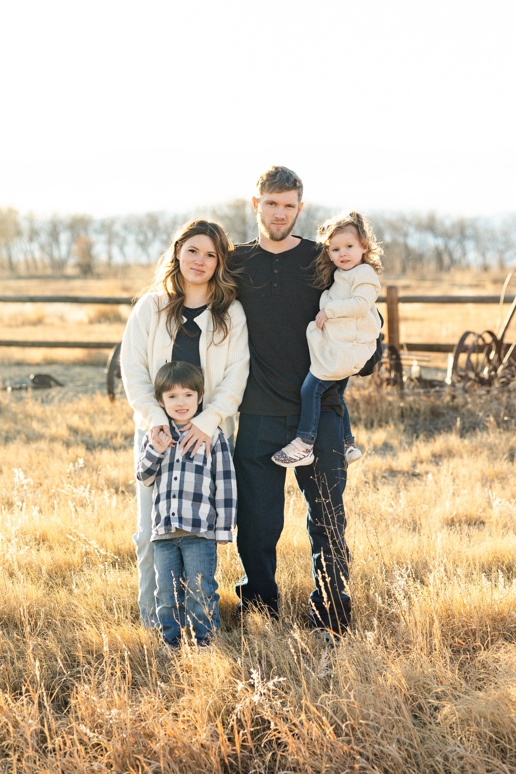 A family of four stands in a field and they smile at the camera.