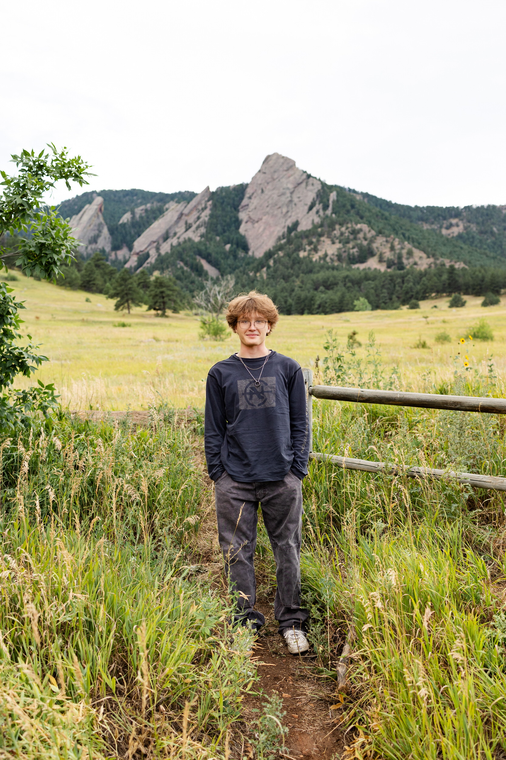 A high school senior boy stands on a mountain trail with his hands in his pockets and smiles at the camera.