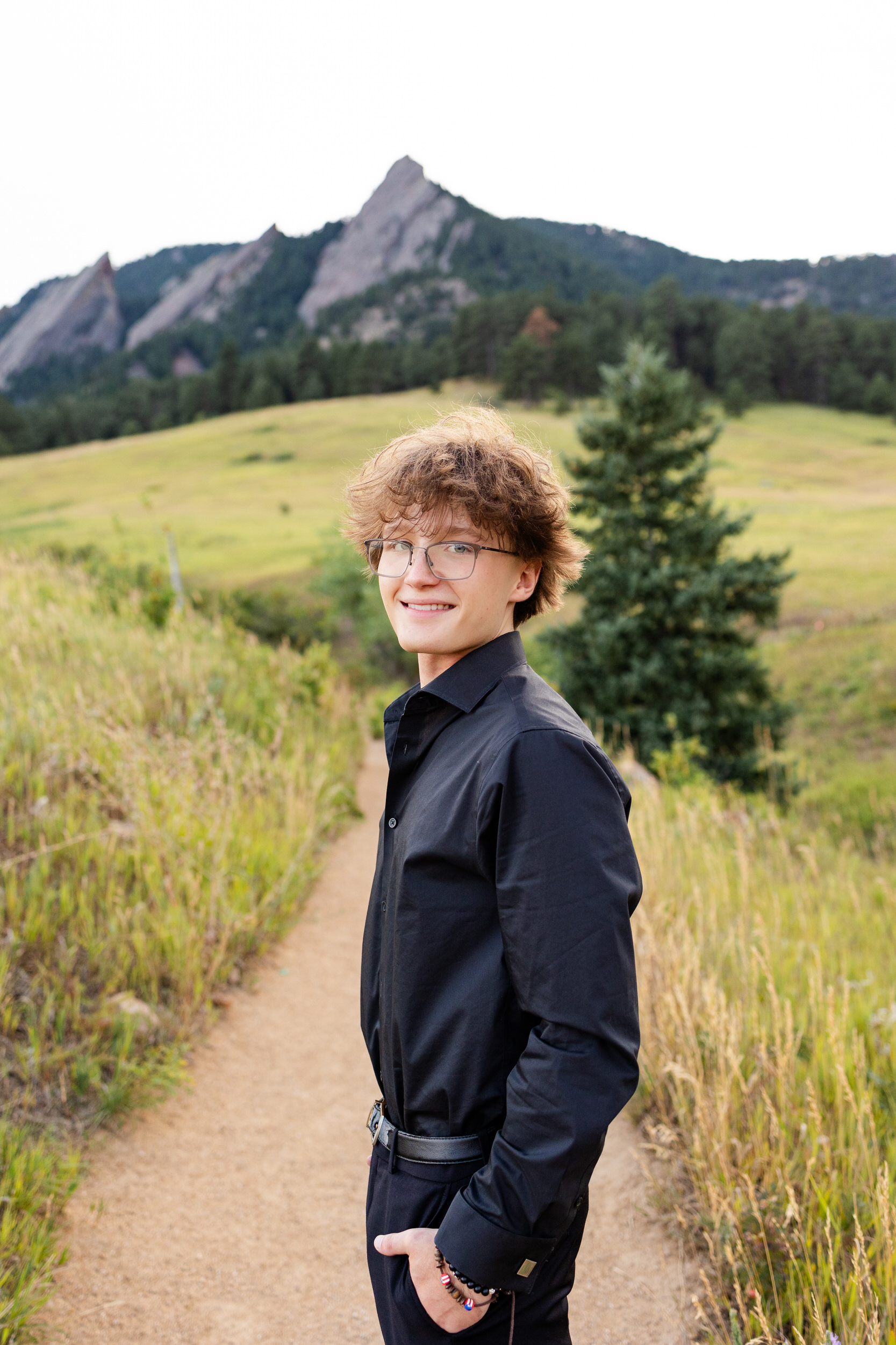A high school senior boy stands on a mountain trail with his hands in his pockets and smiles at the camera.
