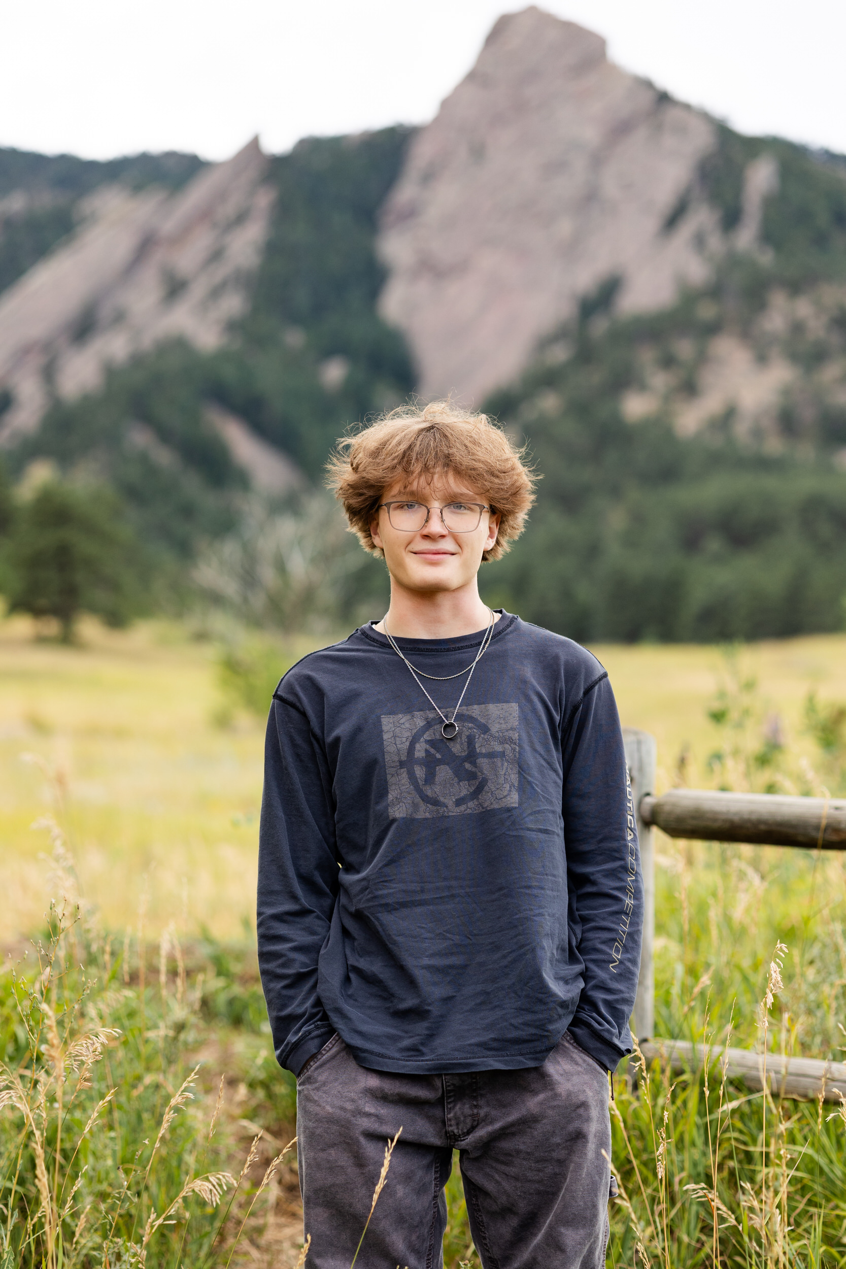 A high school senior boy stands with his hands in his pockets and smiles at the camera with a mountain in the background.