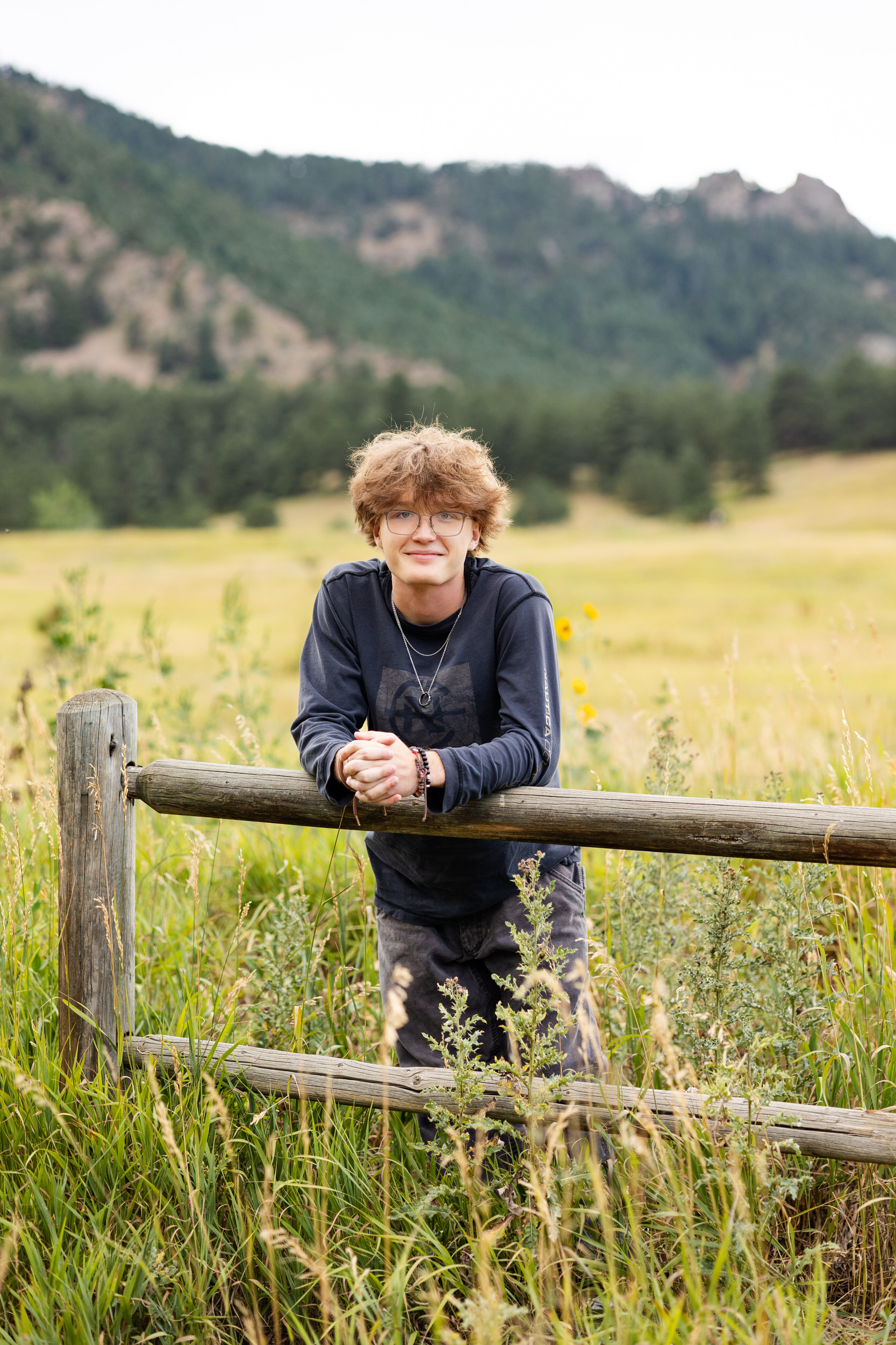 A high school senior boy leans forward on a trailside fence and smiles at the camera.