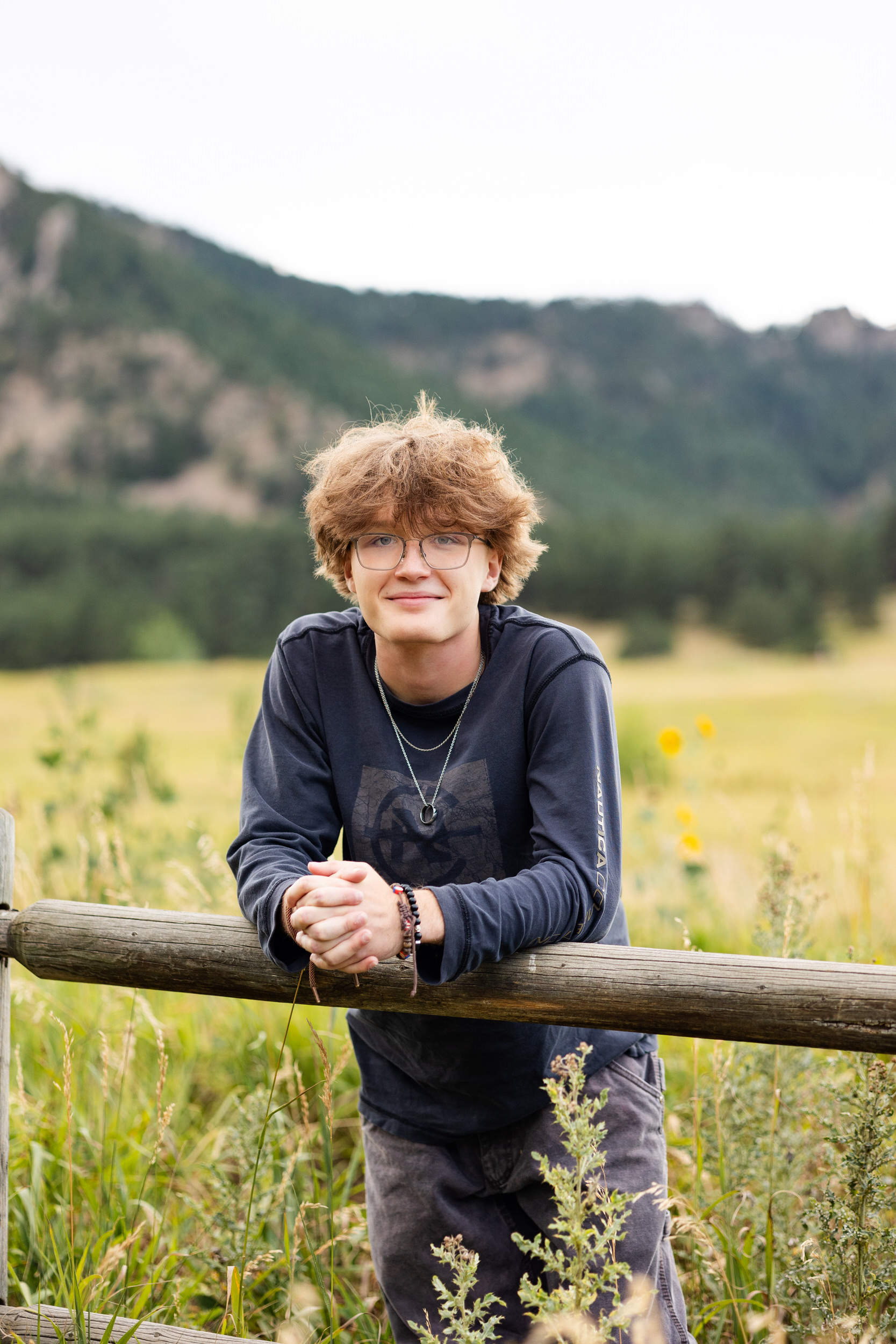 A high school senior boy leans forward on a trailside fence and smiles at the camera.