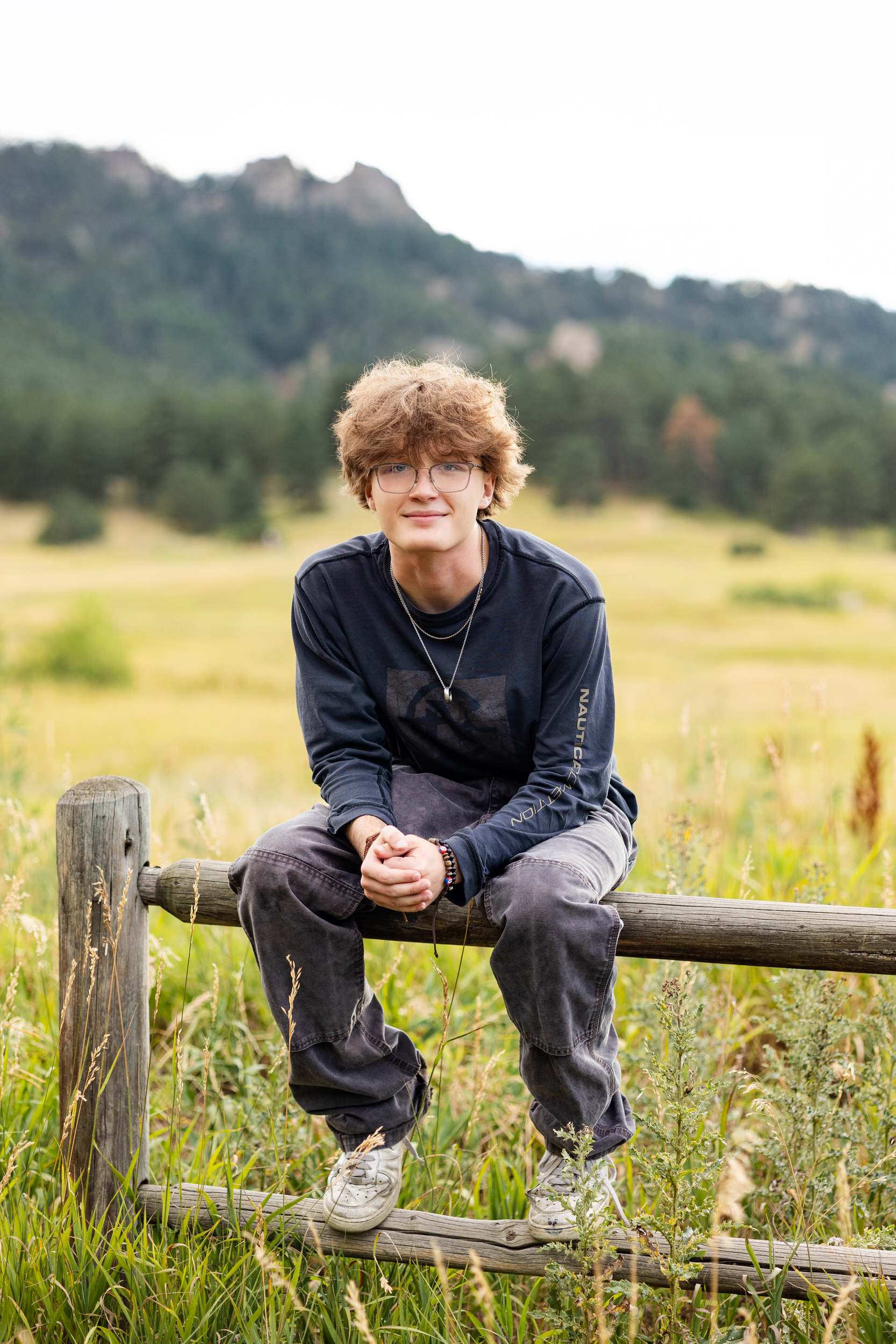 A high school senior boy sits on a trailside fence and smiles at the camera.