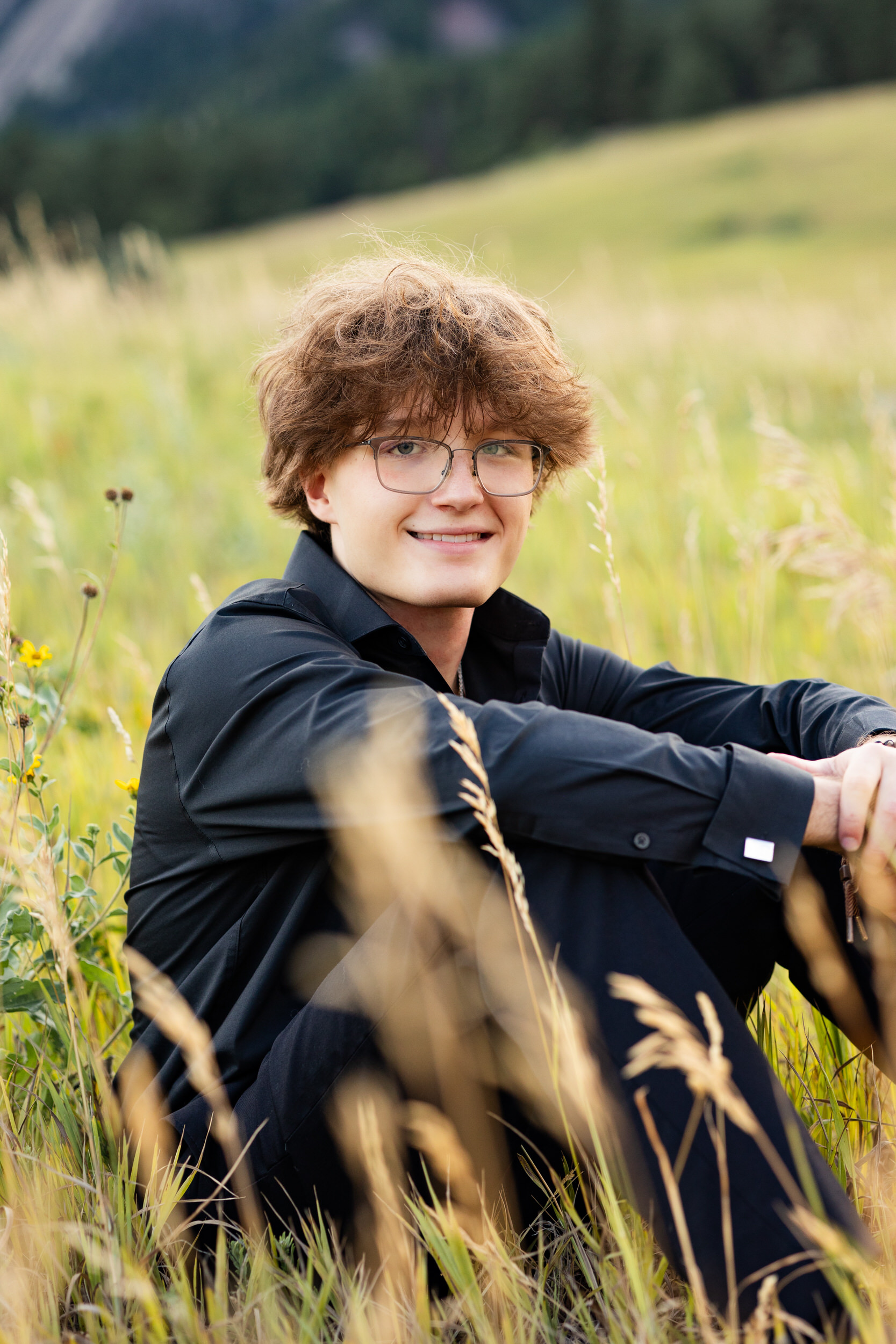 A high school senior boys sits in a field with his arms resting on his knees and smiles at the camera.