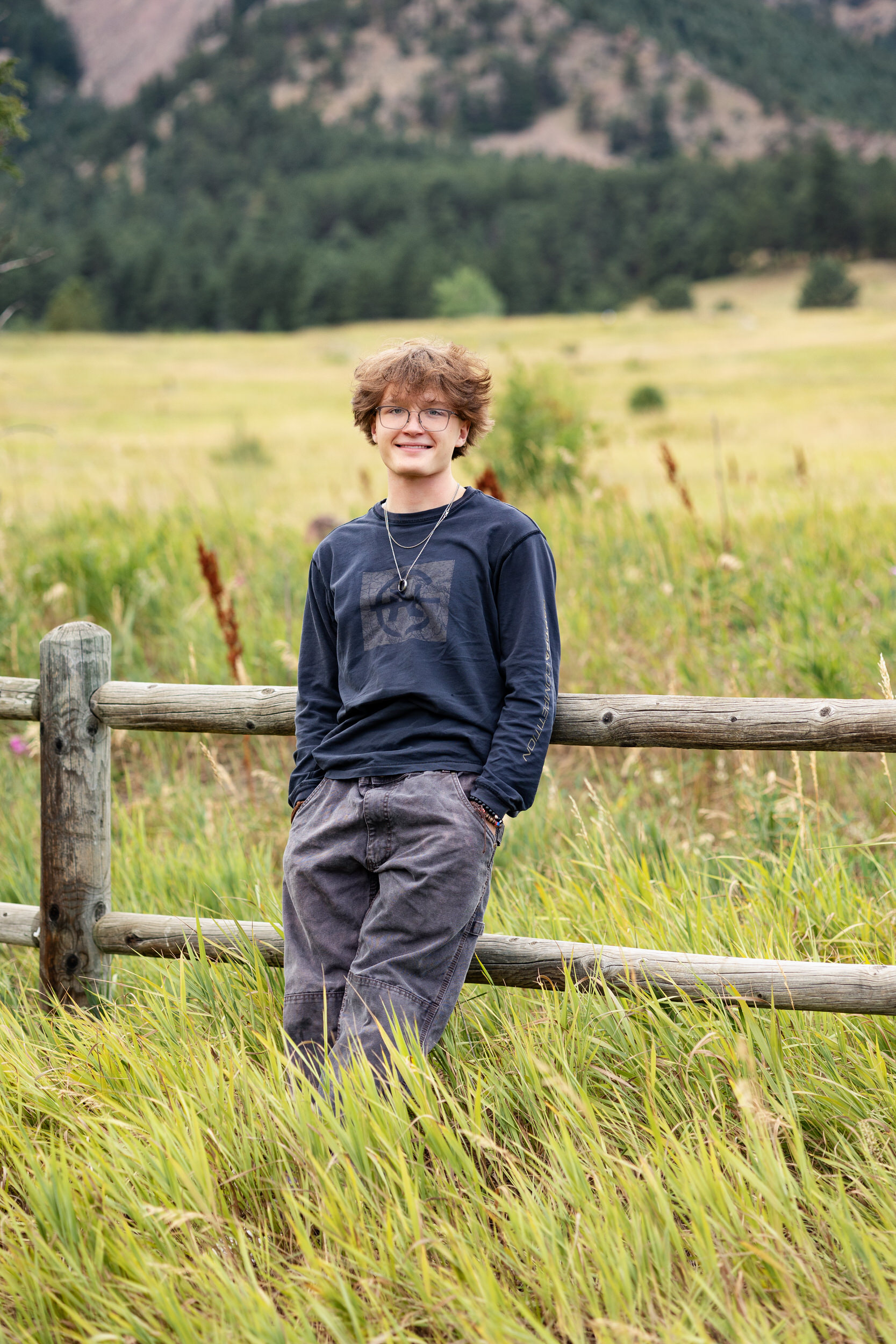 A high school senior boy leans against a trailside fence with his hands in his pockets and smiles at the camera.