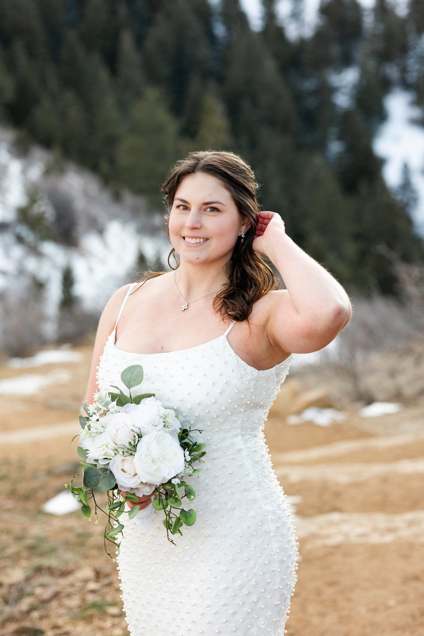 A woman in a white dress holds a bouquet of flowers and runs one hand through her hair and smiles at the camera.