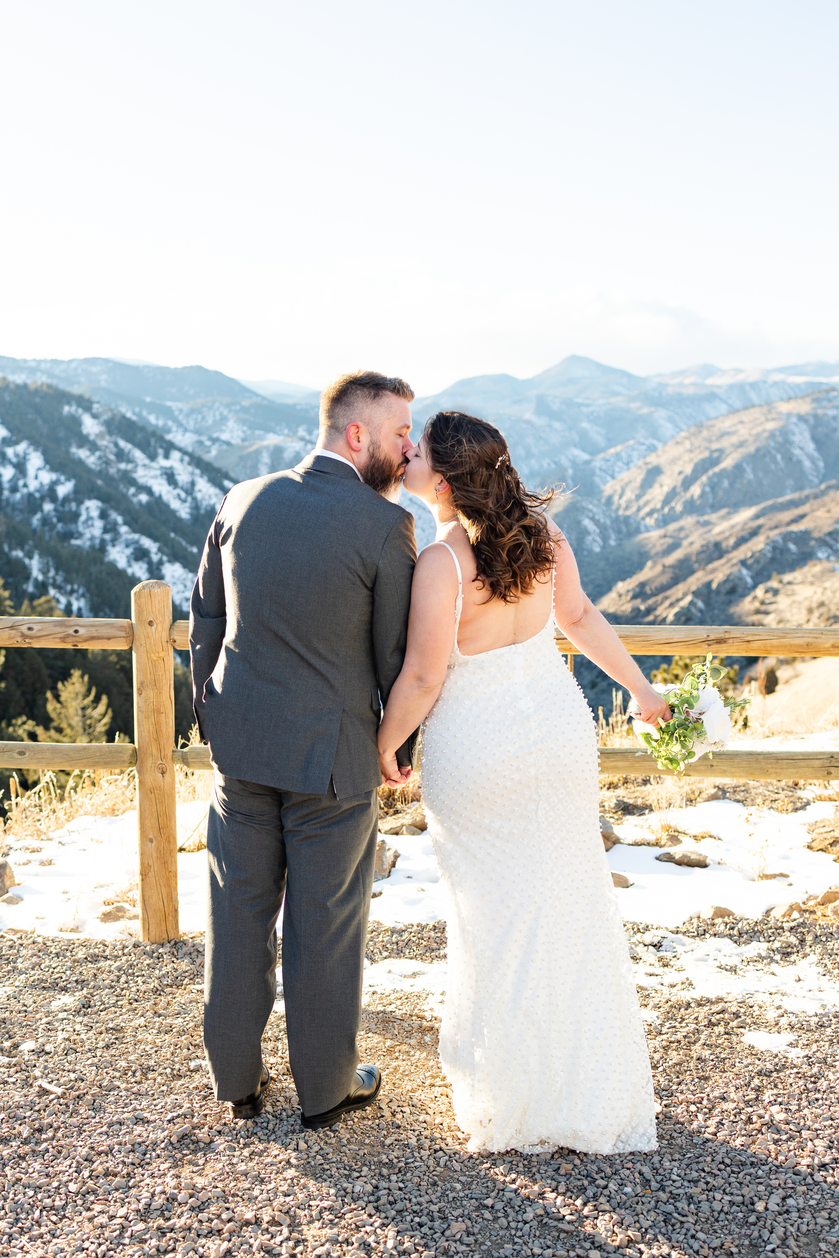A husband and wife in wedding attire stand in front of a grand view of the mountains and share a kiss.