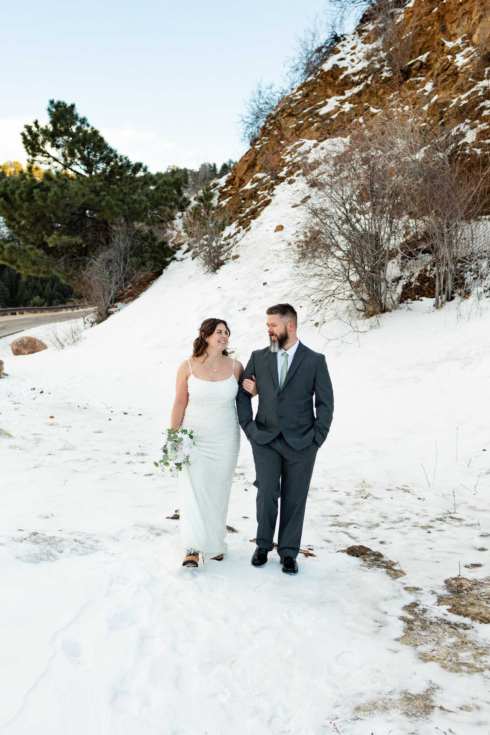 A husband and wife in wedding attire walk through the snow arm-in-arm and smile at each other.