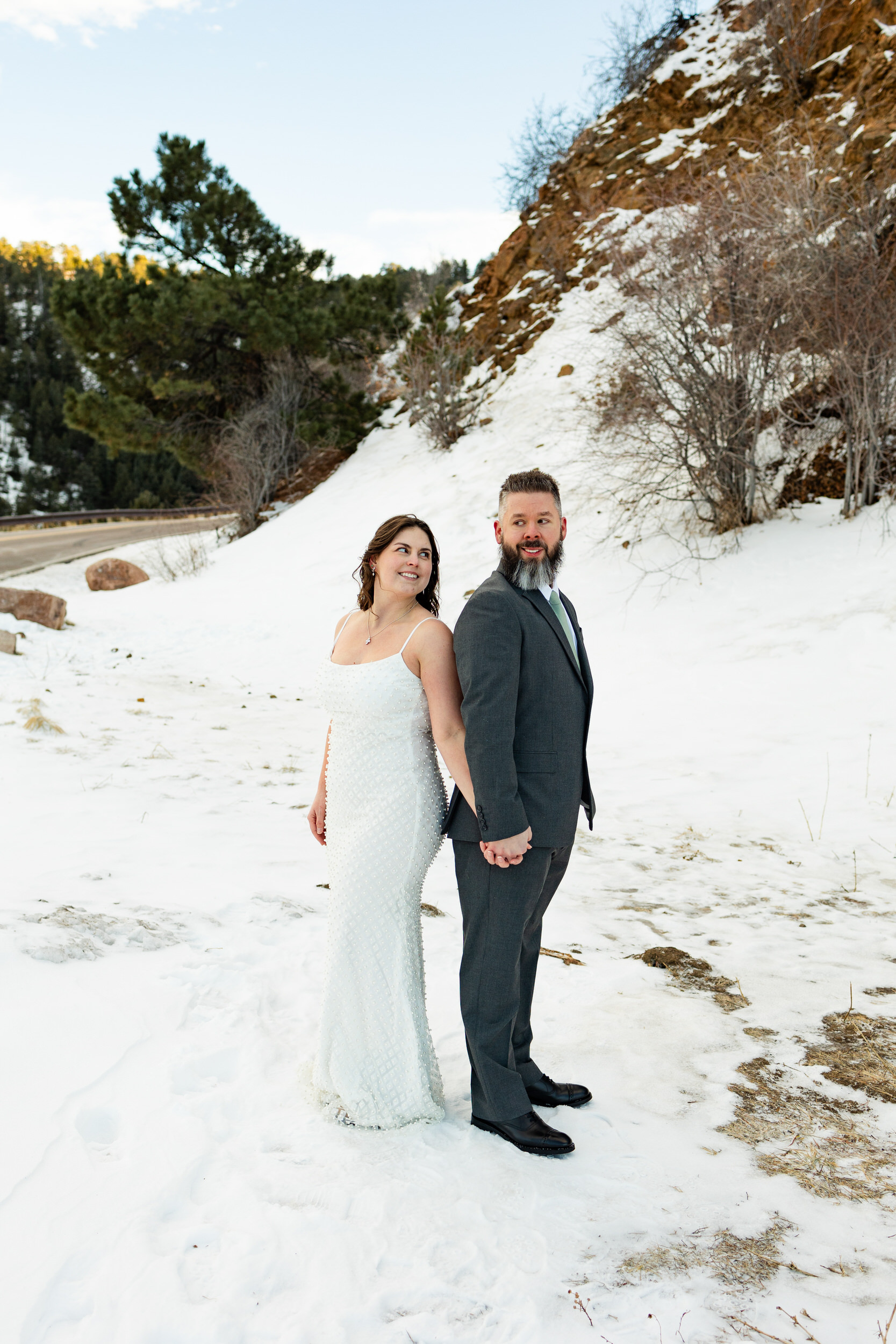 A husband and wife in wedding attire stand back-to-back in the snow and look over their shoulders at each other.