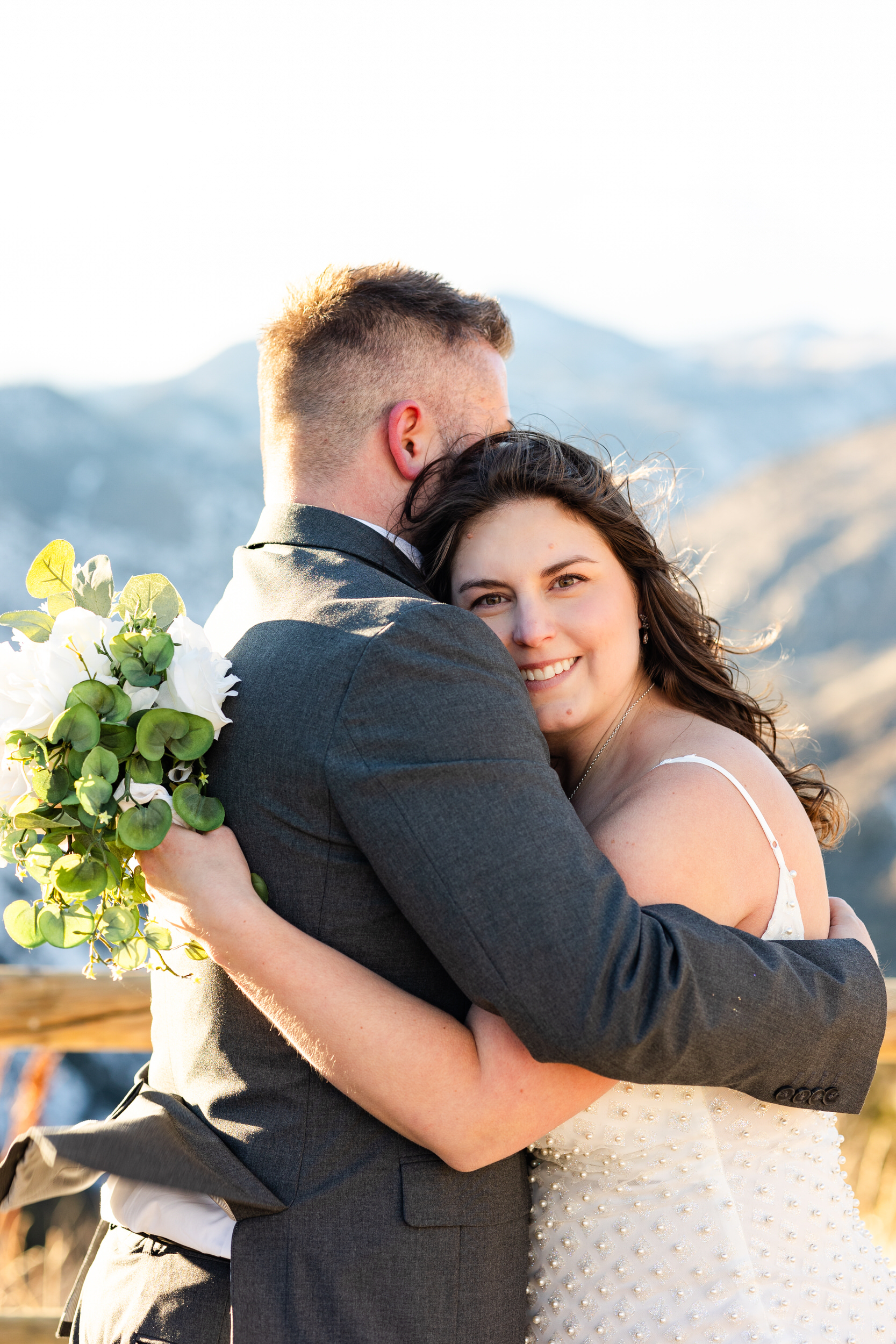 A woman hugs her husband, smiles at the camera while she rests her head on his shoulder, and holds a bouquet of flowers.