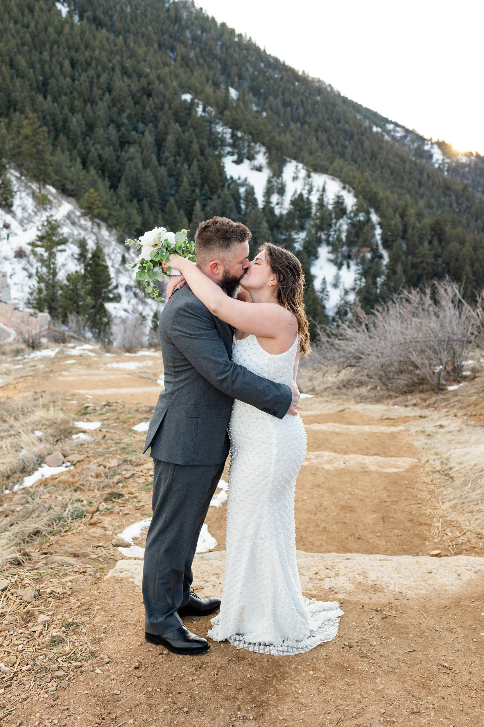 A husband and wife in wedding attire share a kiss in front of the mountains.