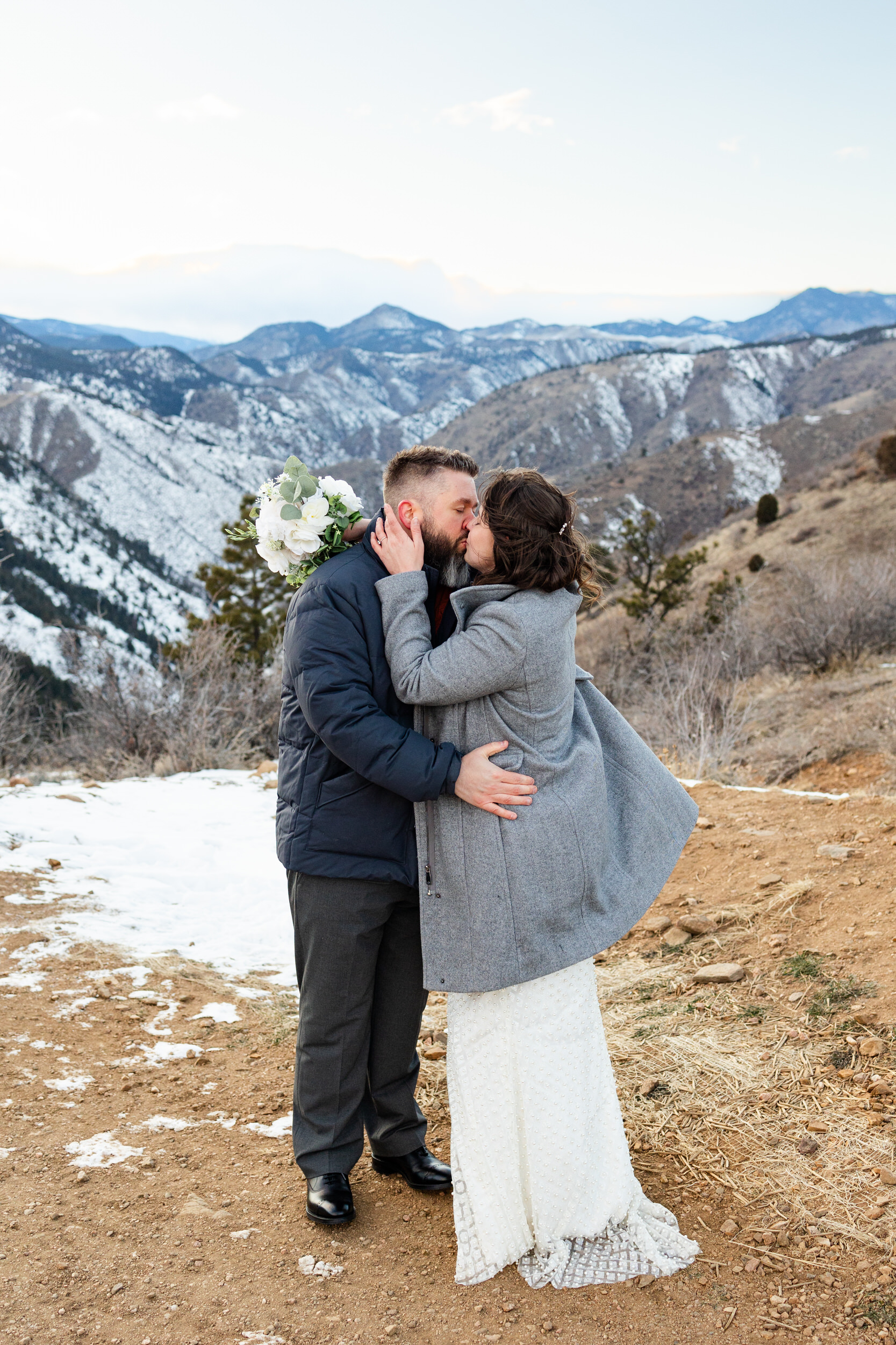 A husband and wife in wedding attire and big coats share a kiss in front of snow-capped mountains.