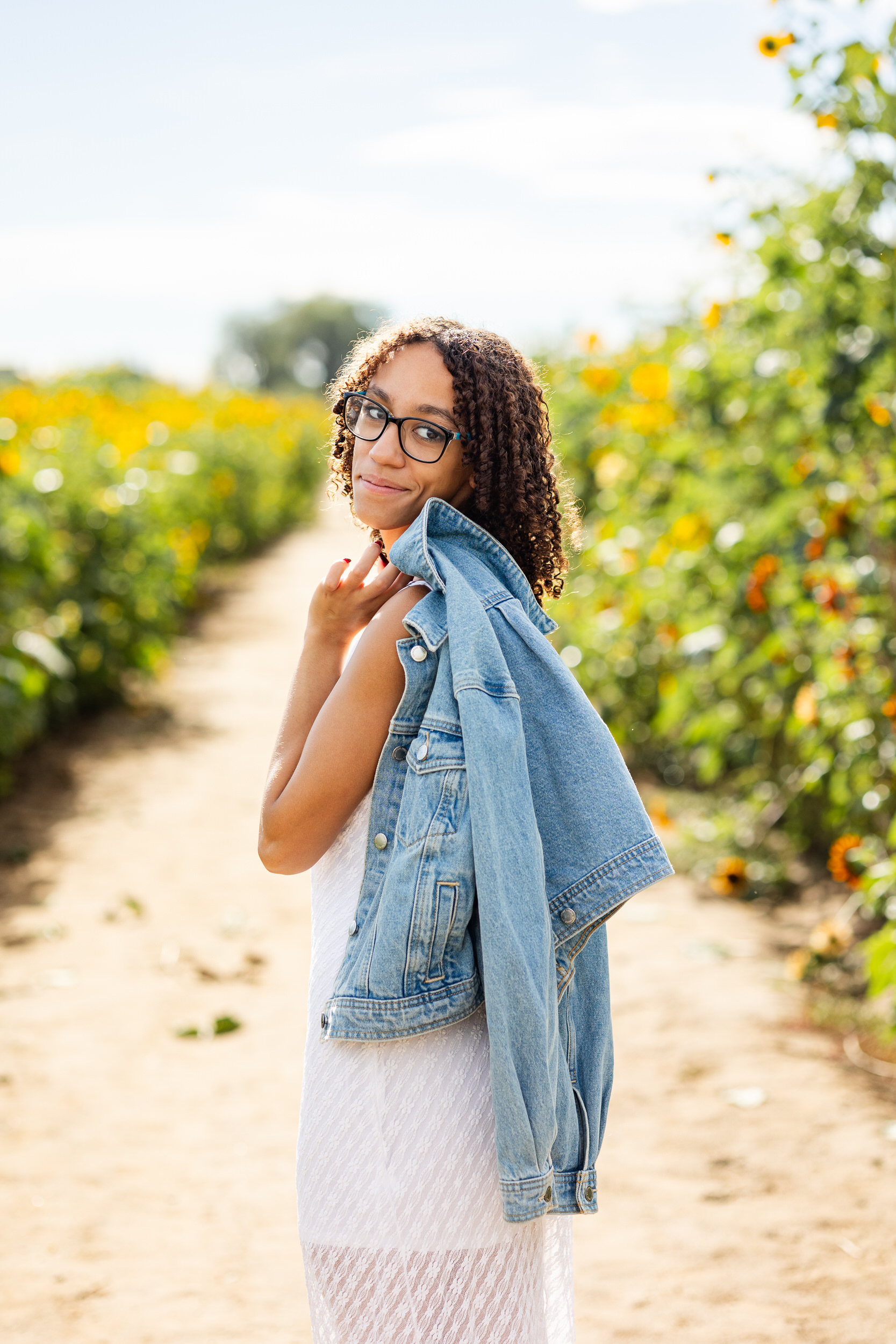A woman in a white dress throws a denim jacket over her shoulder and smiles at the camera.