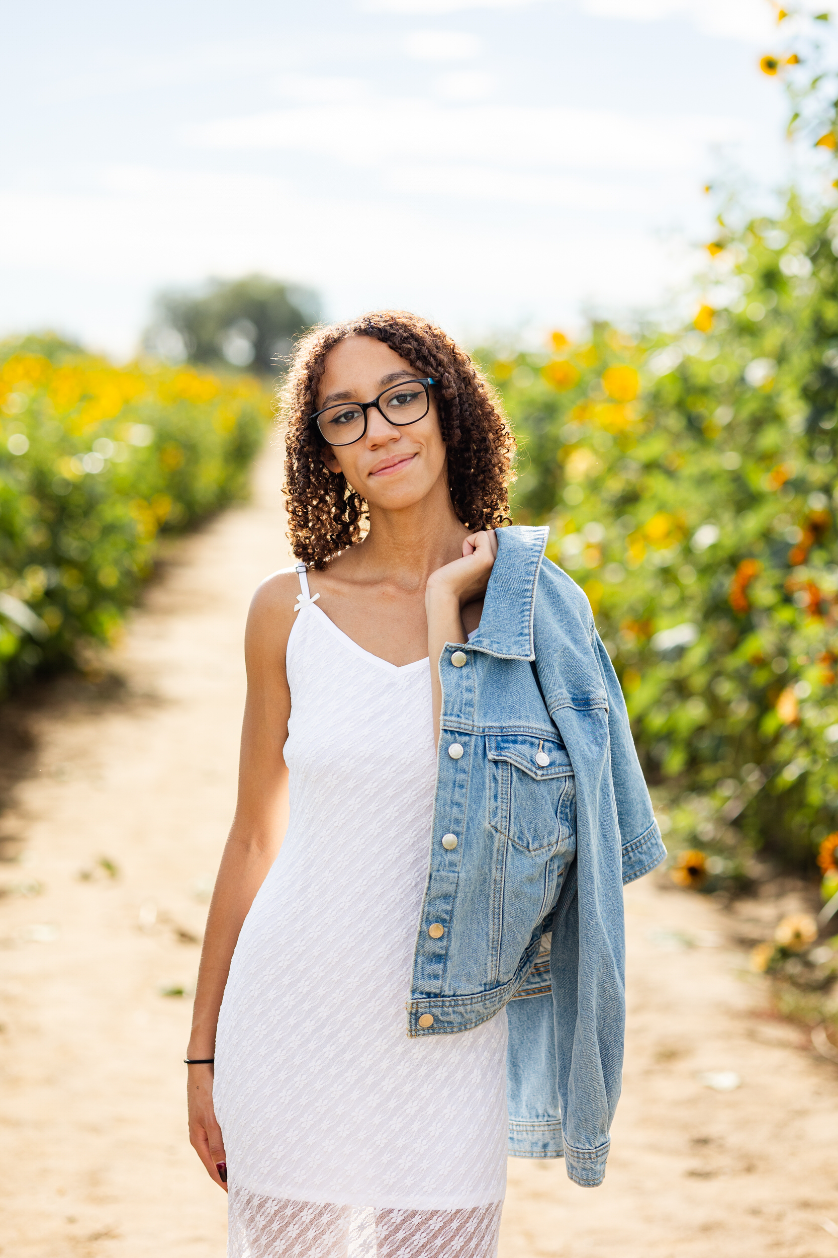 A woman in a white dress throws a denim jacket over her shoulder and smiles at the camera.