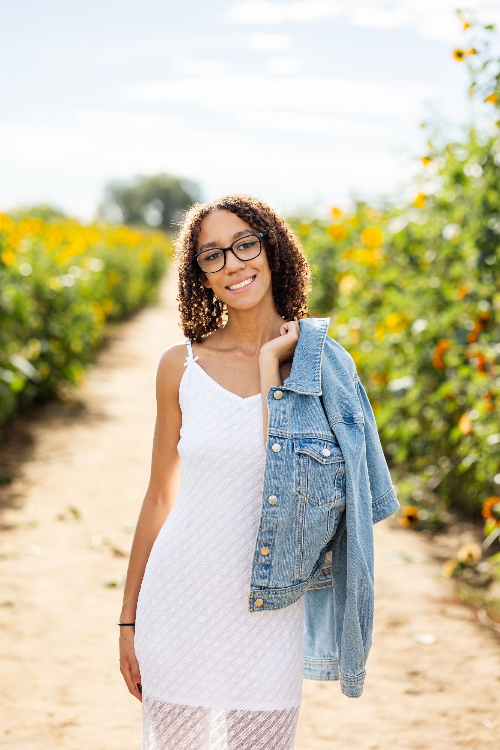 A woman in a white dress throws a denim jacket over her shoulder and smiles at the camera.