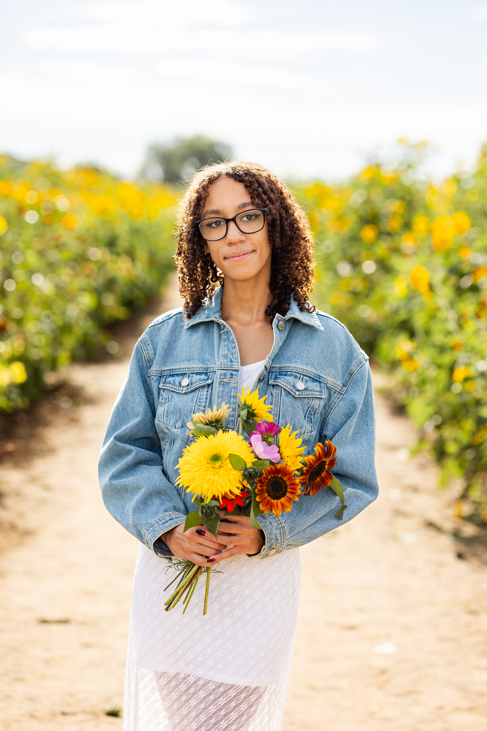 A young woman in a white dress and denim jacket holds a bouquet of sunflowers and smiles at the camera.
