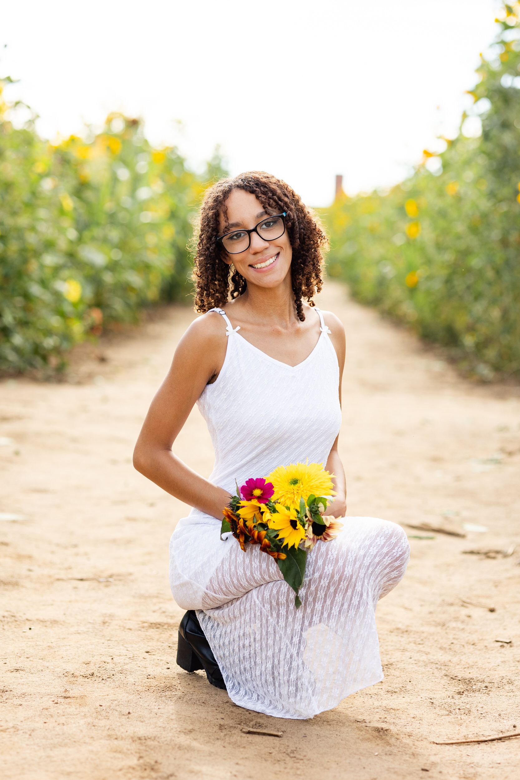 A young woman in a white dress squats down in a sunflower field with a bouquet of sunflowers and smiles at the camera.