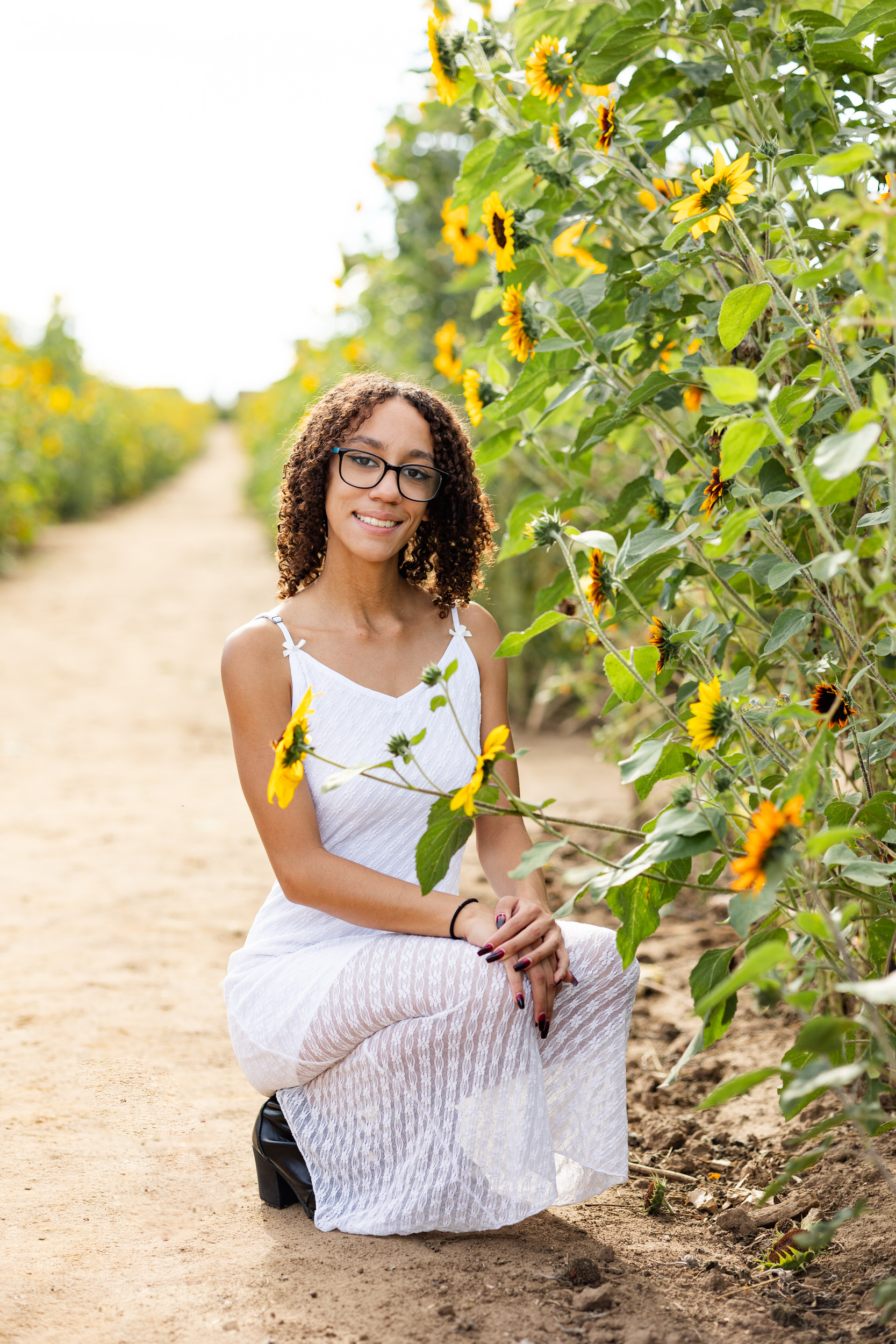A young woman in a white dress squats down next to sunflowers and smiles at the camera.